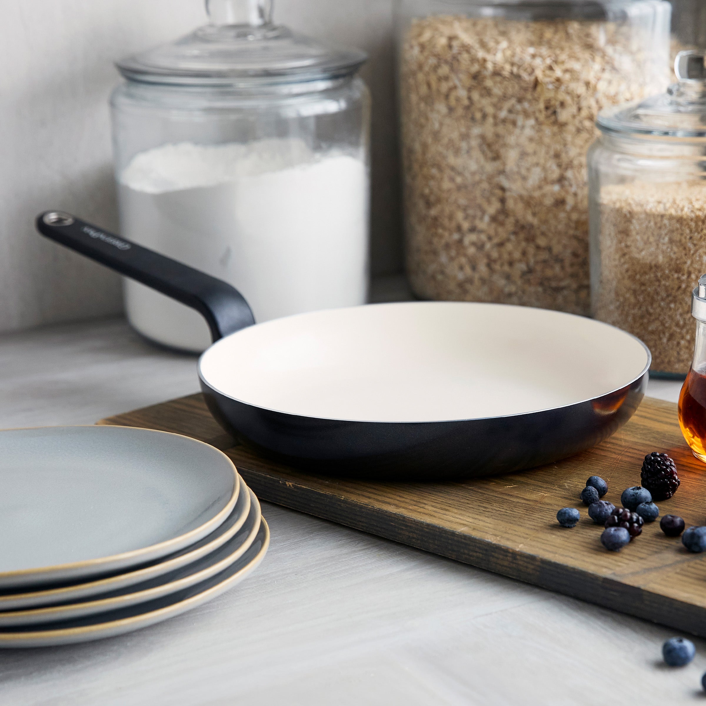 Black ceramic nonstick 12-inch frypan on a wooden board with plates, berries, and jars of flour and oats in the background.