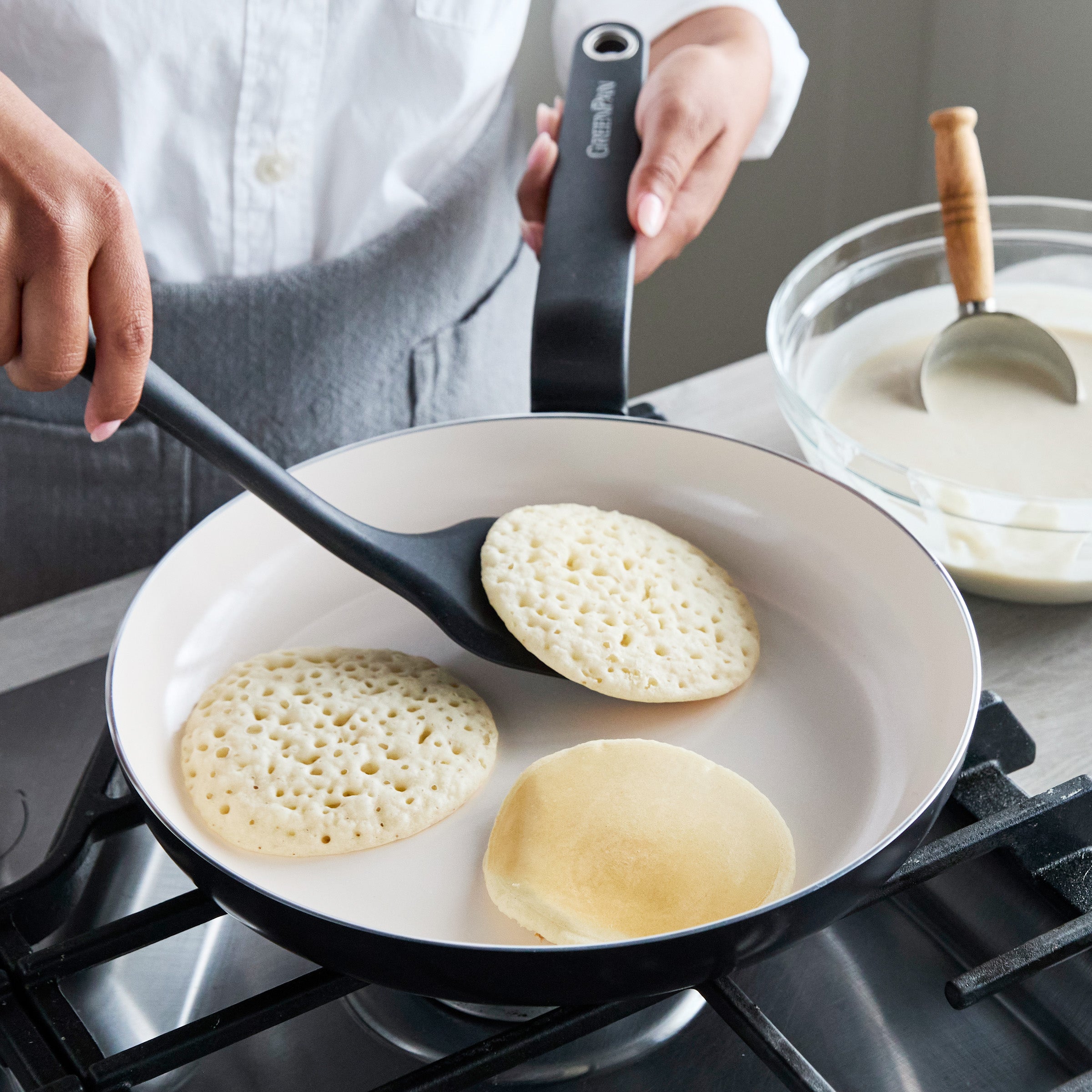 A pancake being flipped in a black ceramic nonstick 12 frypan, with batter and a ladle beside the stove.