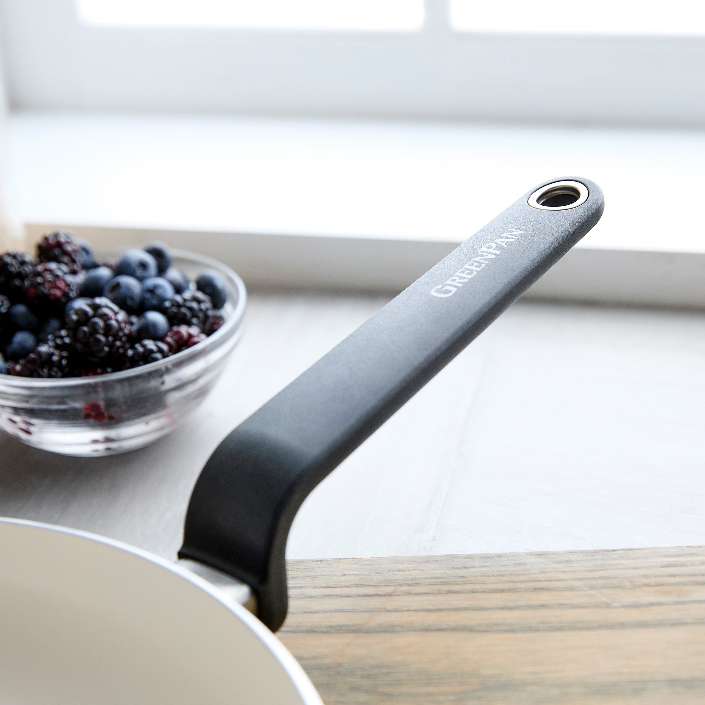 Black ceramic 12-inch frypan with handle on a light kitchen countertop, bowl of berries in the background.