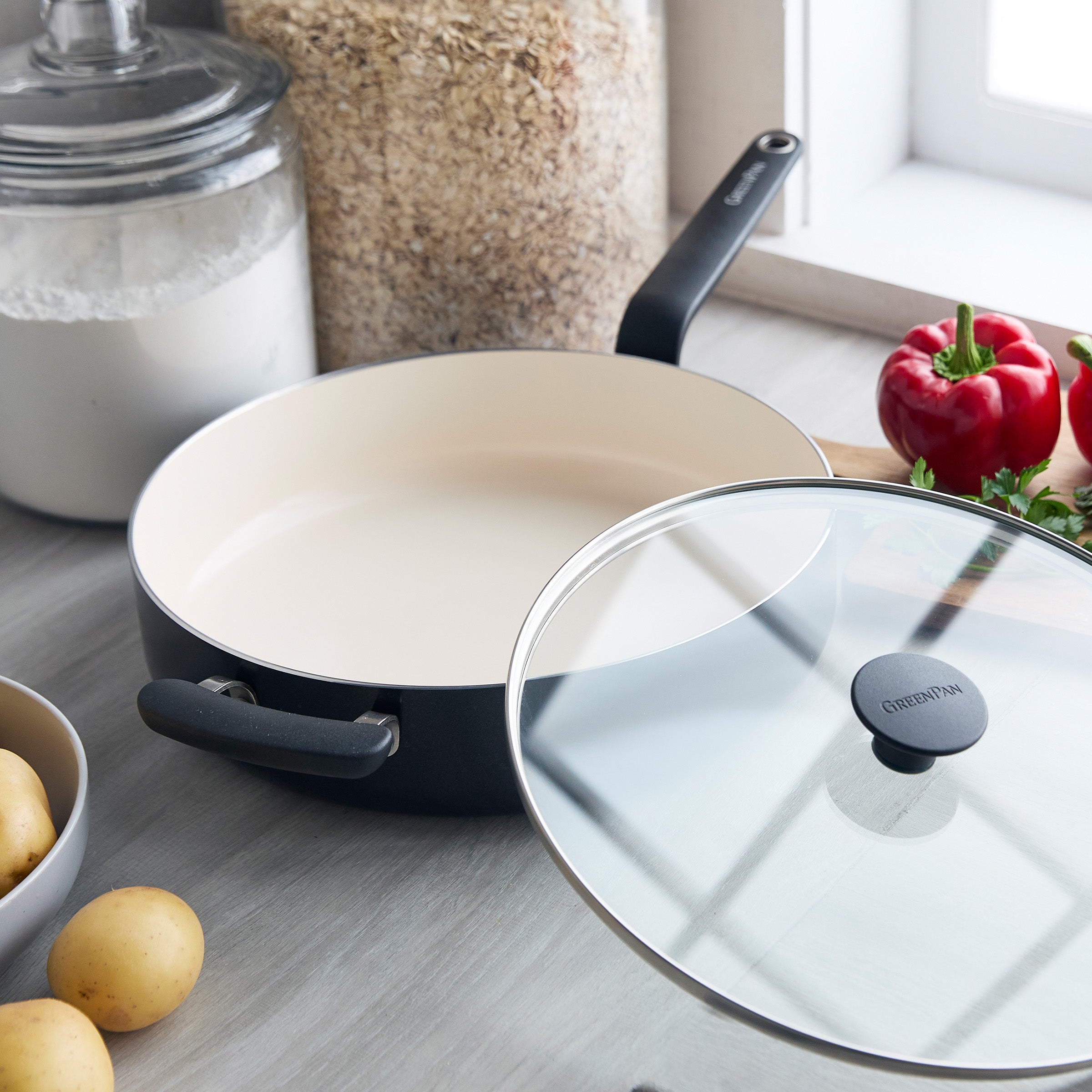 Green ceramic sauté pan with lid on a countertop, surrounded by potatoes, flour, oats, and assorted vegetables.