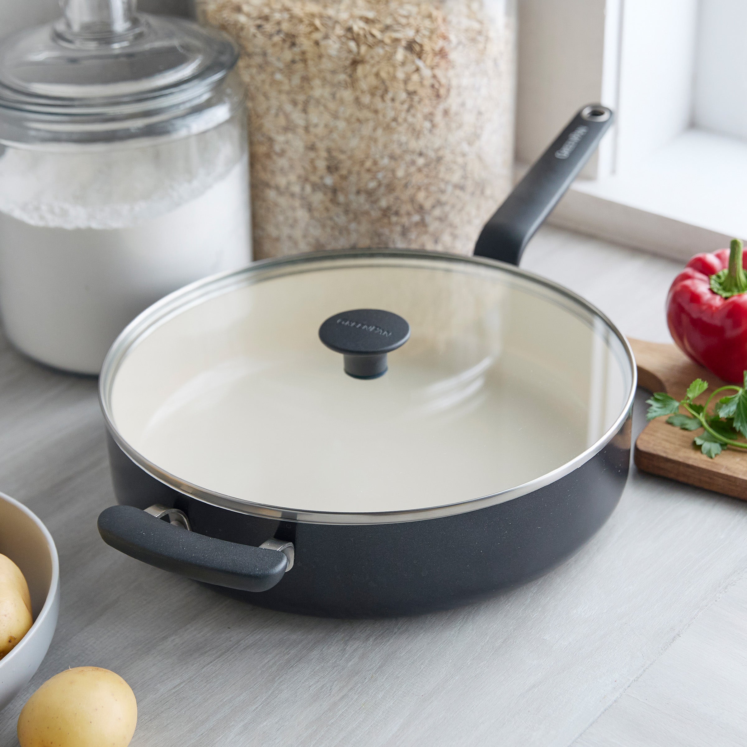 Black ceramic 5-quart sauté pan with lid on a counter next to potatoes, oats, flour, herbs, and a pepper.