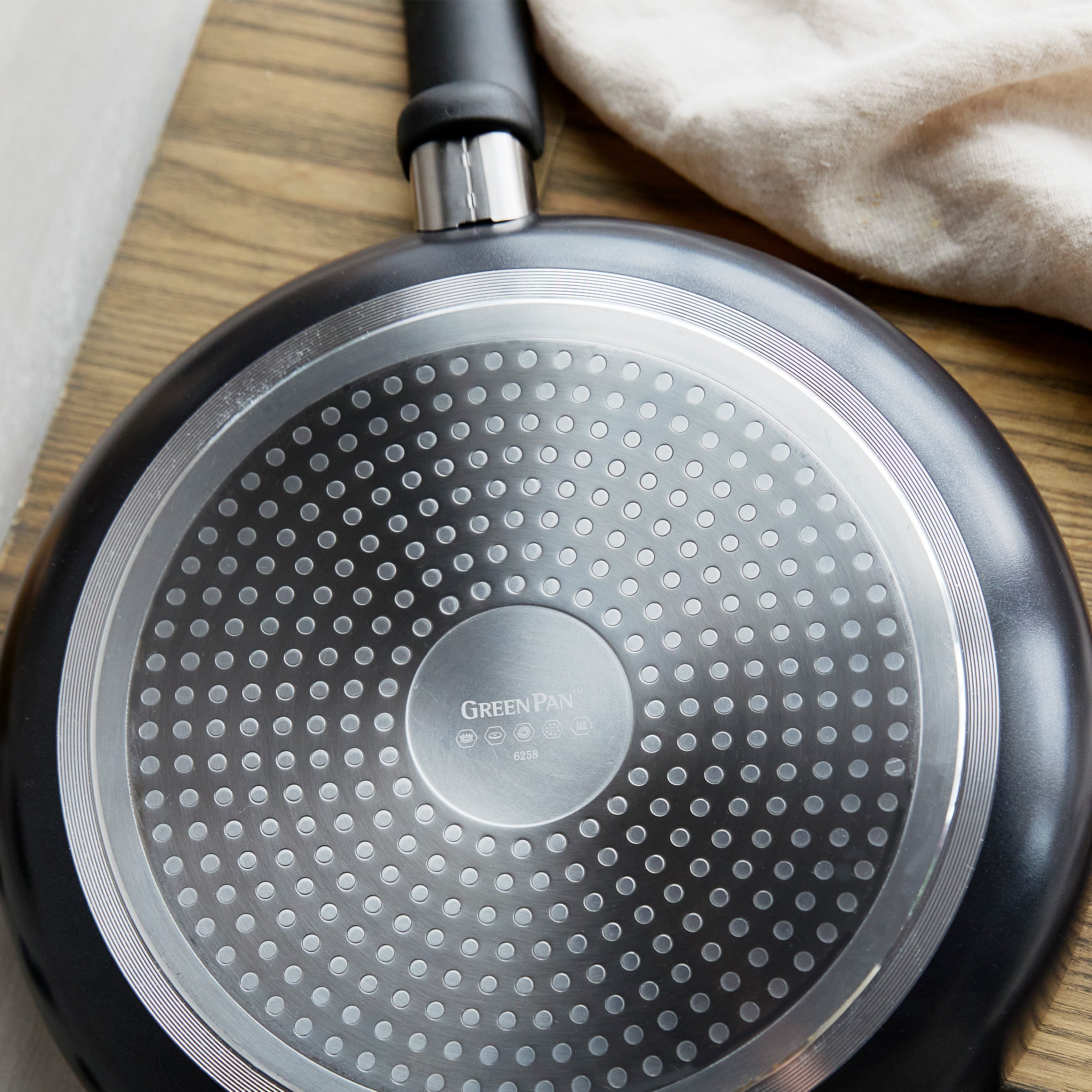Black ceramic nonstick 5-quart sauté pan with lid resting on wood surface next to a light-colored cloth.
