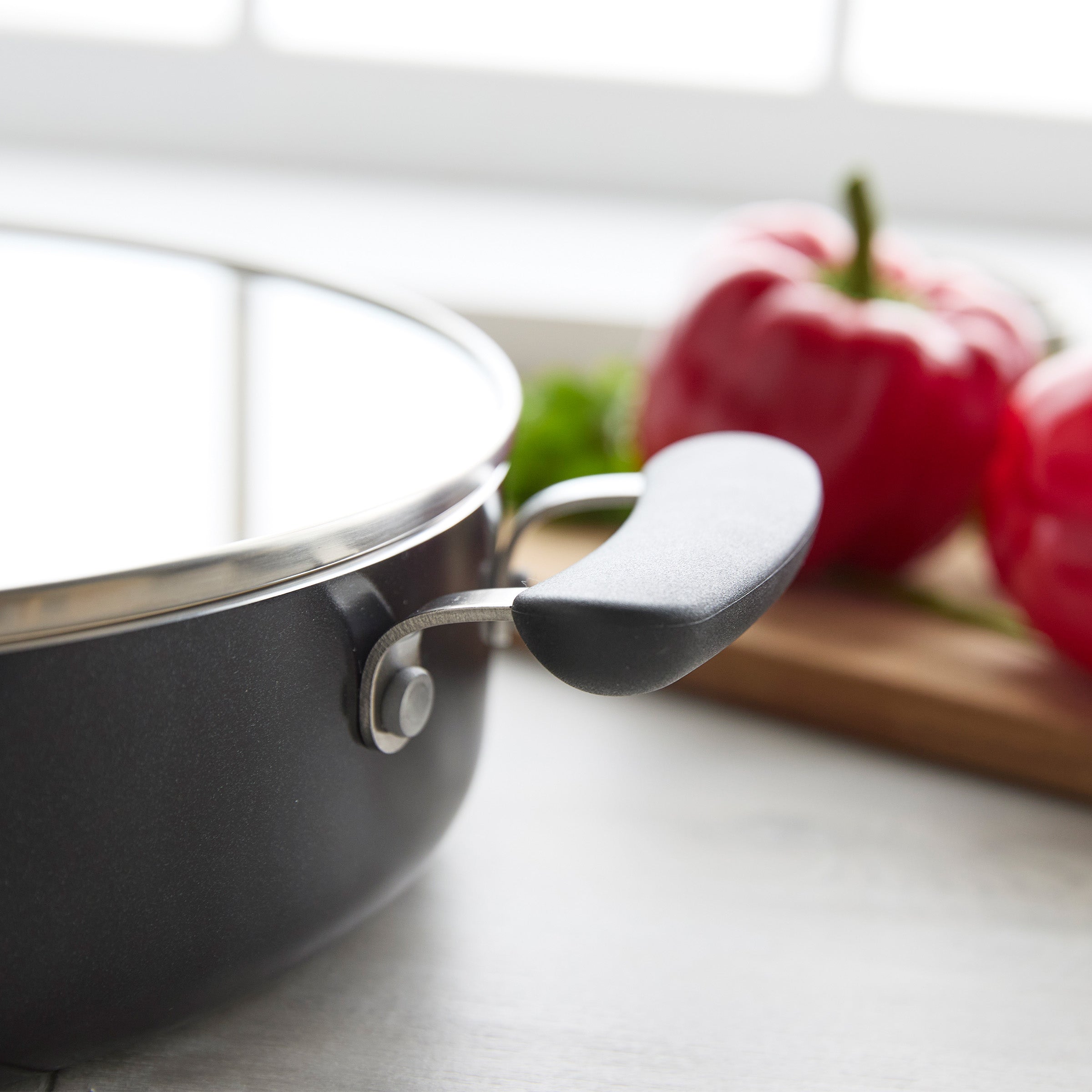 Black ceramic nonstick sauté pan with lid and handle next to red peppers on a wooden cutting board.
