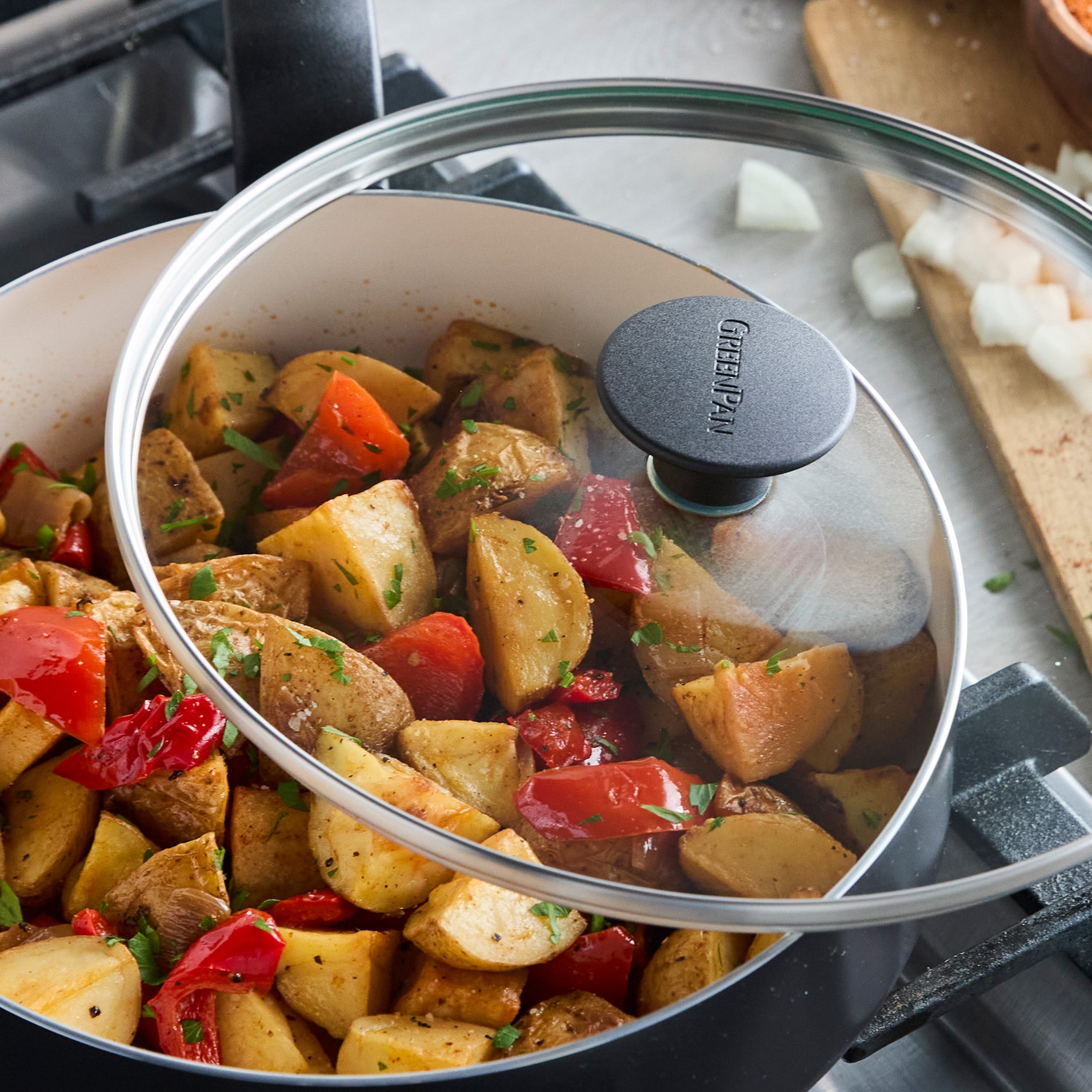 Cubed potatoes and peppers cooking in a black ceramic sauté pan with glass lid; chopped onions on the counter nearby.