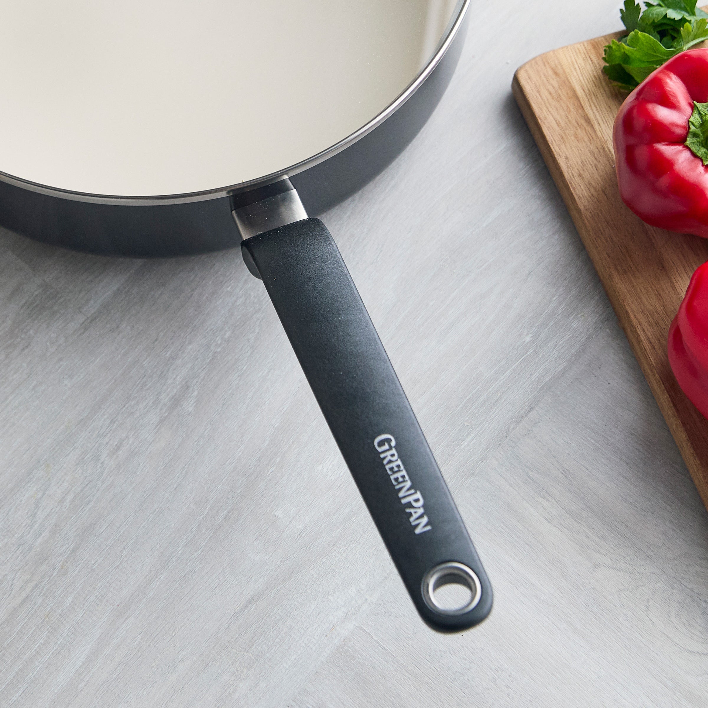 Black 5-quart ceramic nonstick sauté pan with lid on a light counter, next to a board holding peppers and parsley.