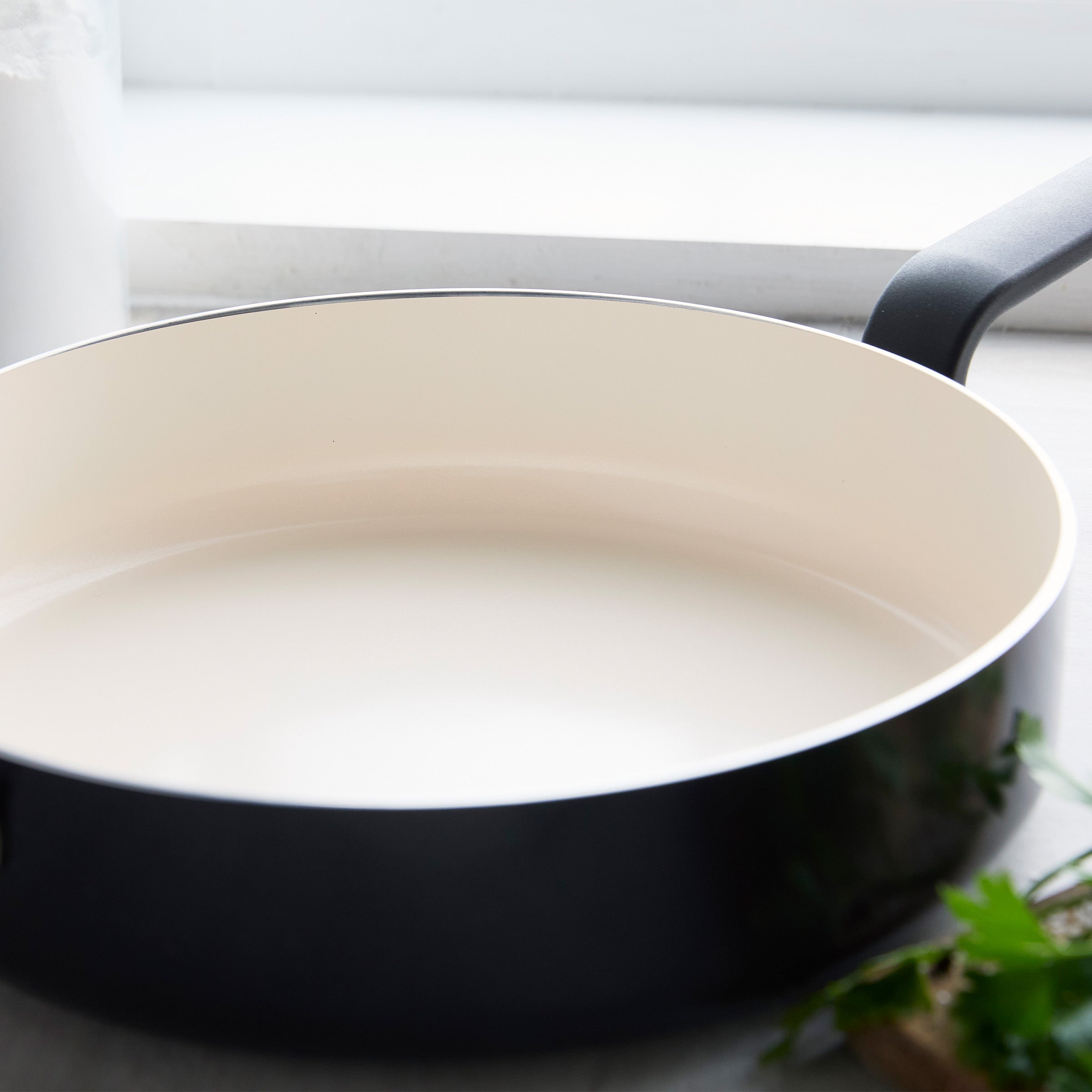 Black ceramic nonstick 5-quart sauté pan with lid on a light kitchen surface, fresh herbs nearby.