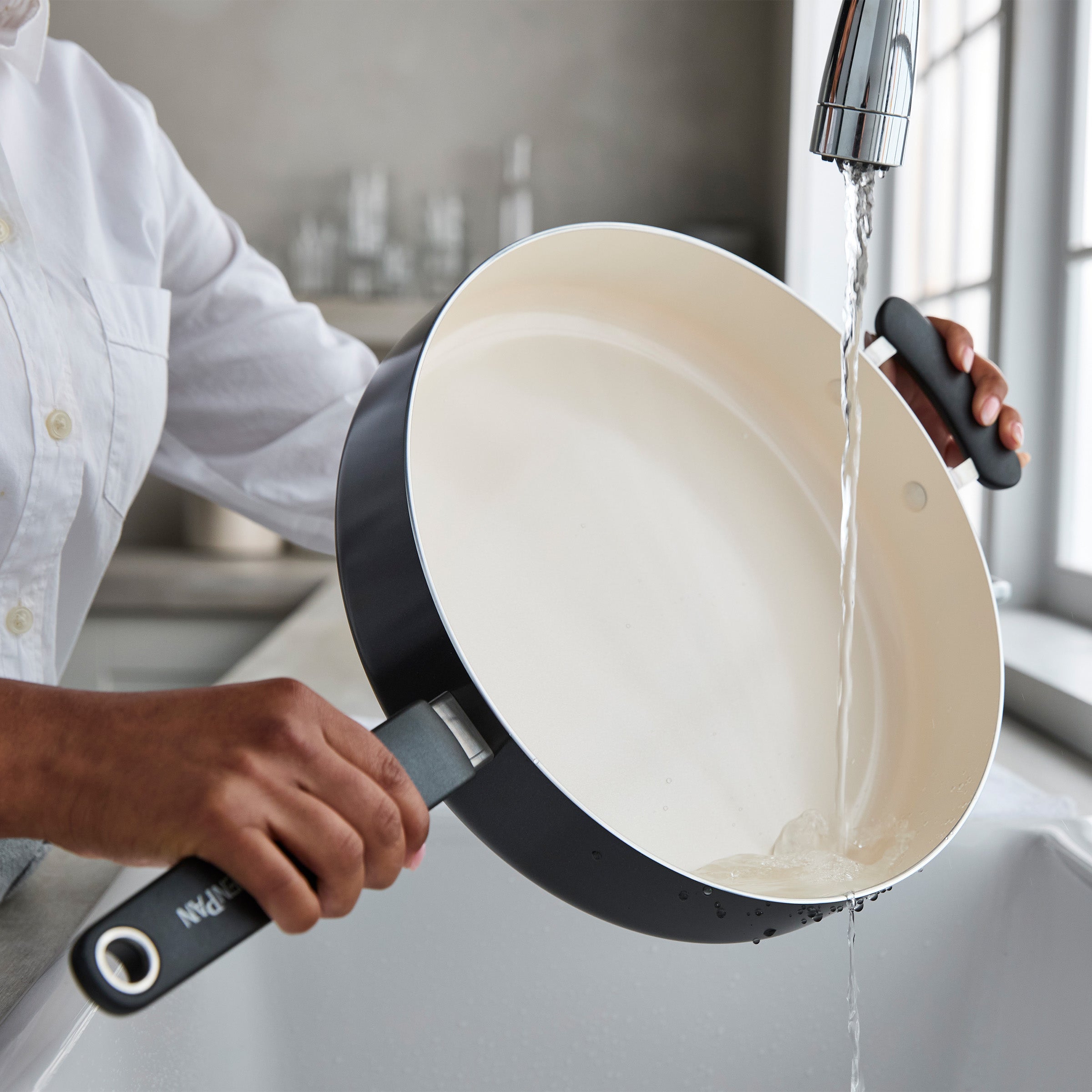 Black ceramic nonstick sauté pan with lid being rinsed under running water in a kitchen sink.