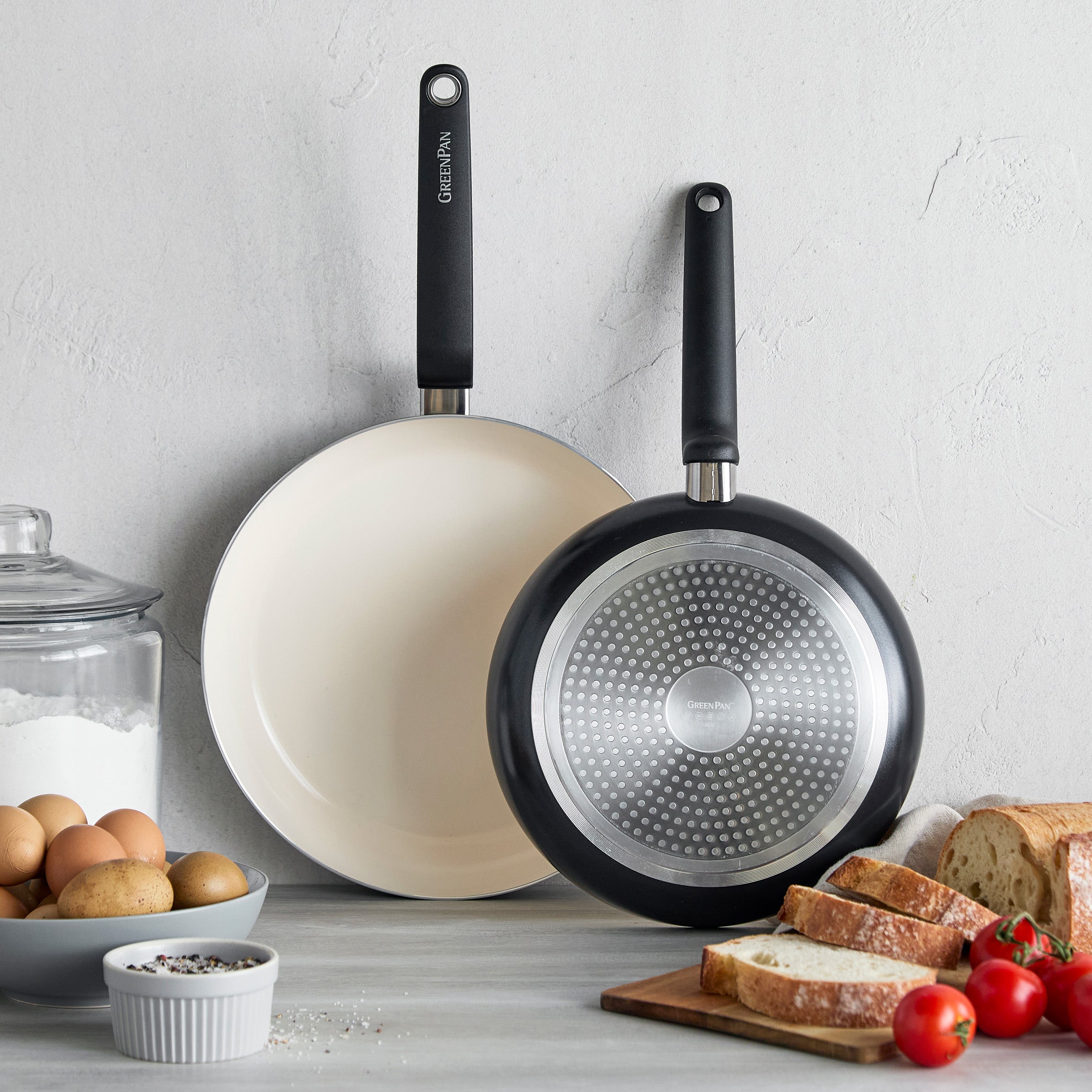 Two black ceramic nonstick frypans, 10 and 12, on a counter near bread, tomatoes, eggs, and a salt shaker.