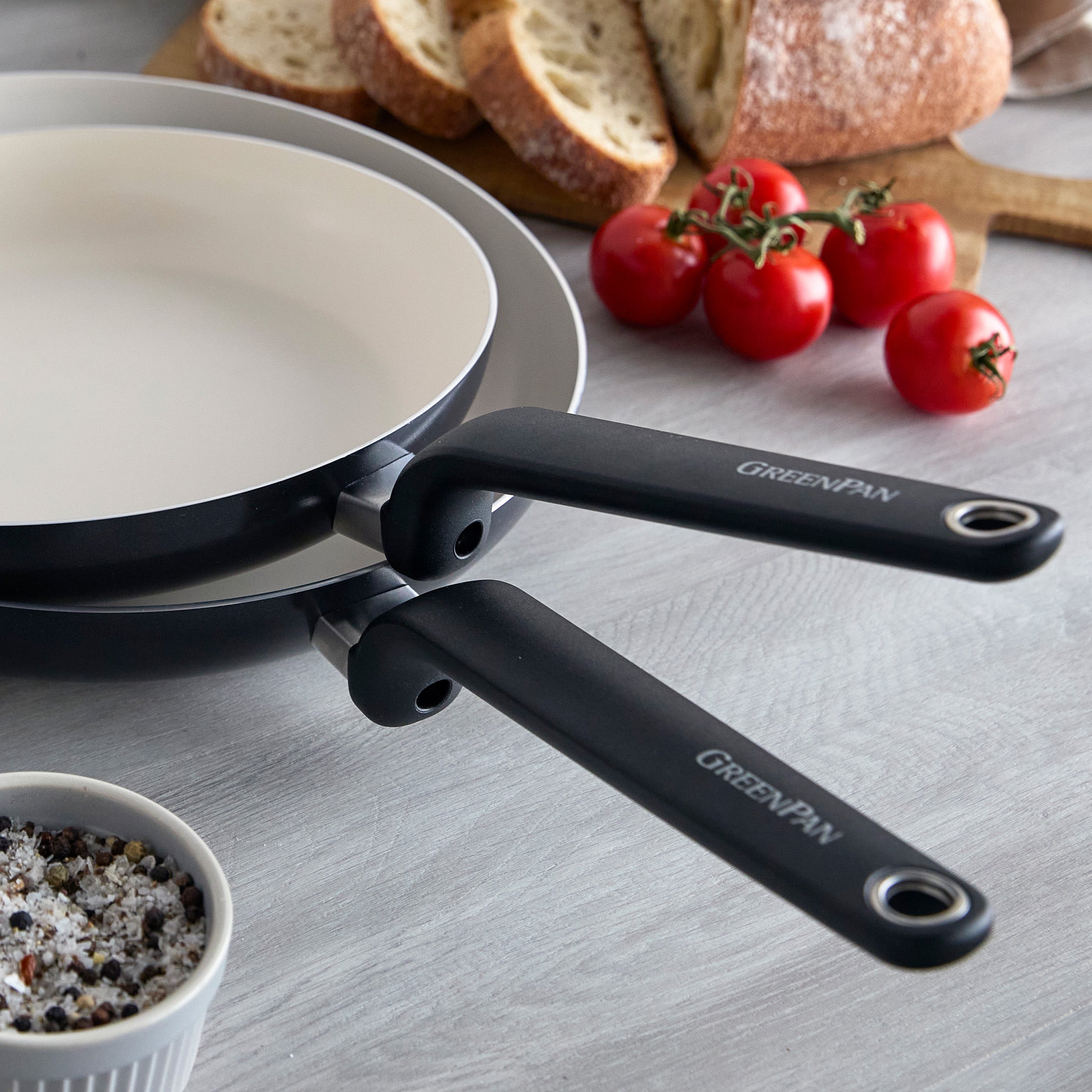 Two black ceramic nonstick frypans, 10 and 12, stacked on a kitchen counter with tomatoes and bread nearby.