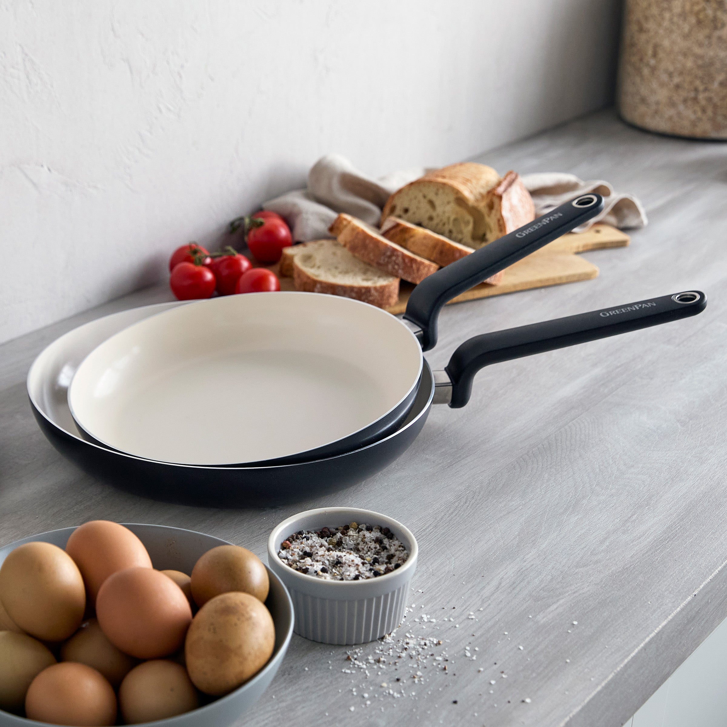 Set of two black ceramic nonstick frypans, 10 and 12, on a counter with eggs, bread, tomatoes, and seasonings.
