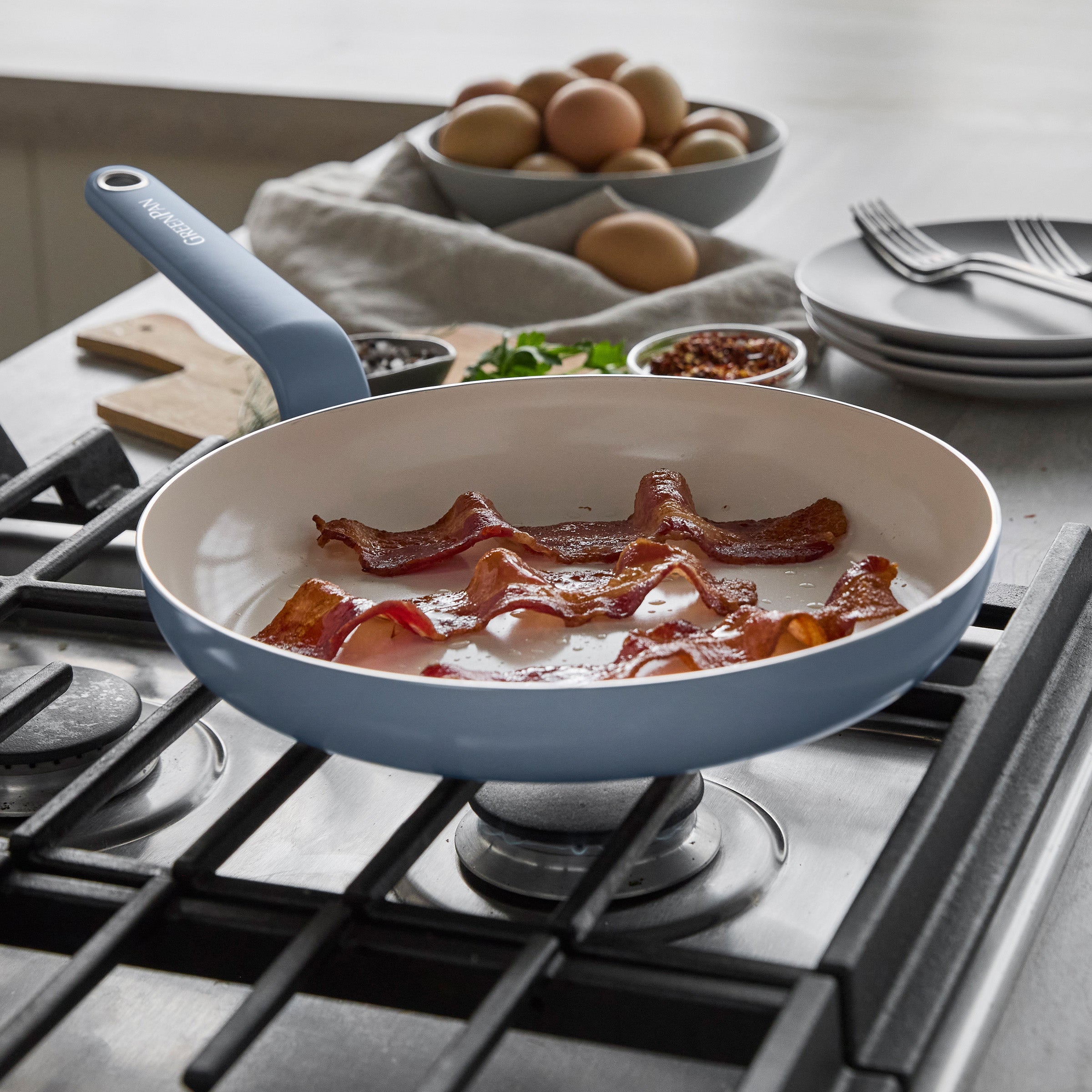 Bacon cooking in a blue ceramic nonstick frypan set, with eggs, plates, and seasonings visible in the background.