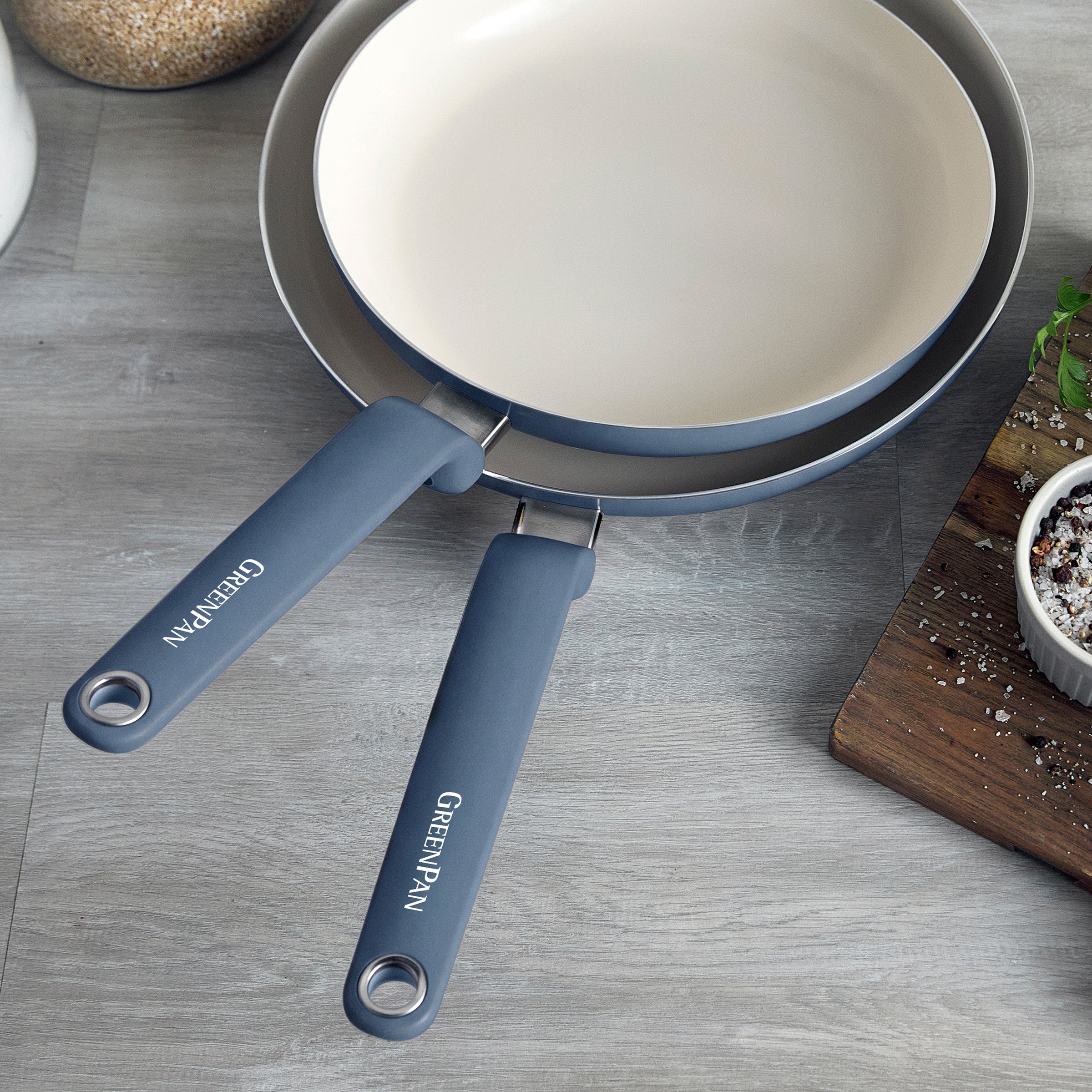 Two Dutch blue ceramic frypans, 10 and 12, stacked on a grey surface next to a cutting board sprinkled with spices.