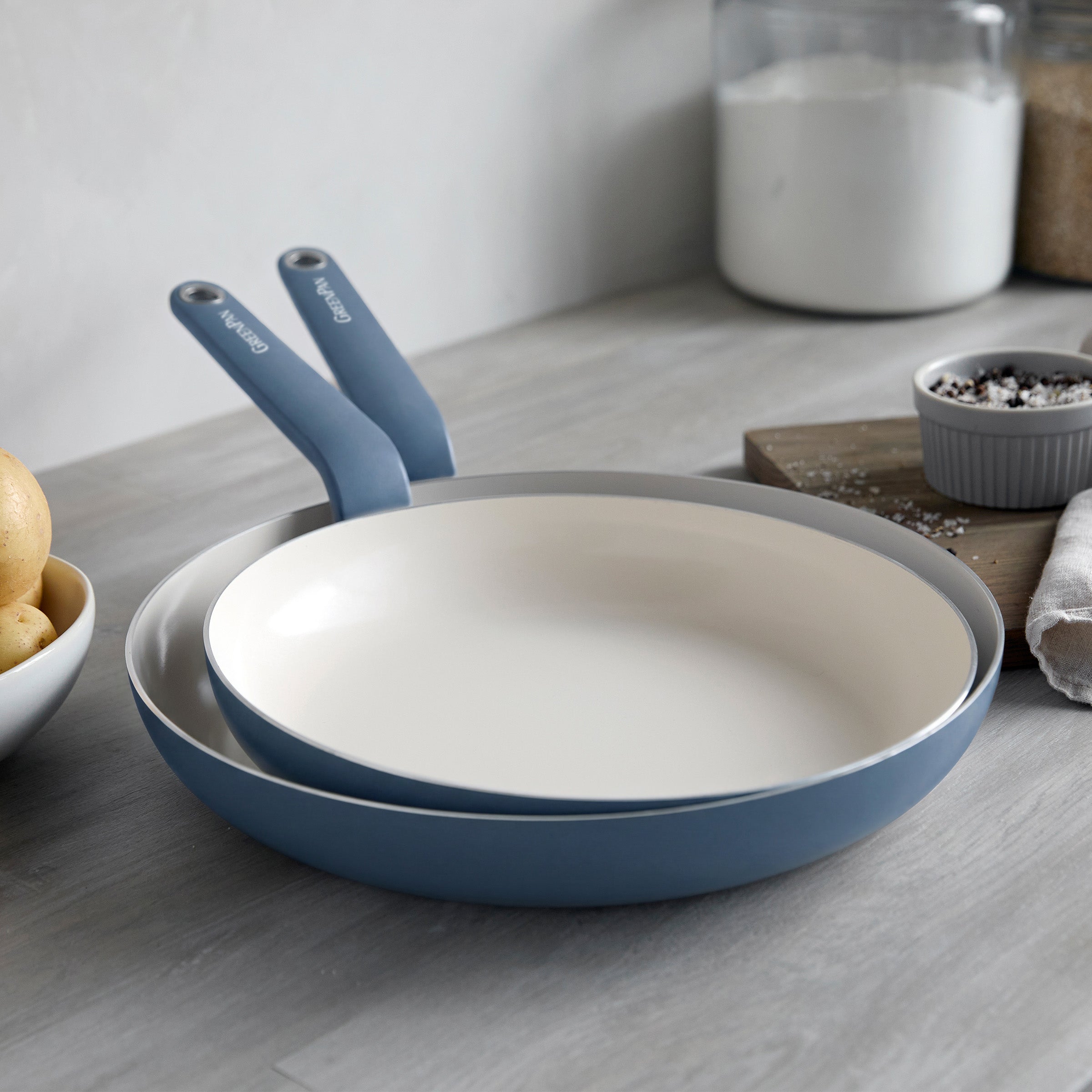 Two Dutch blue ceramic nonstick frypans, 10 and 12, on a gray counter with jars and a bowl of potatoes nearby.