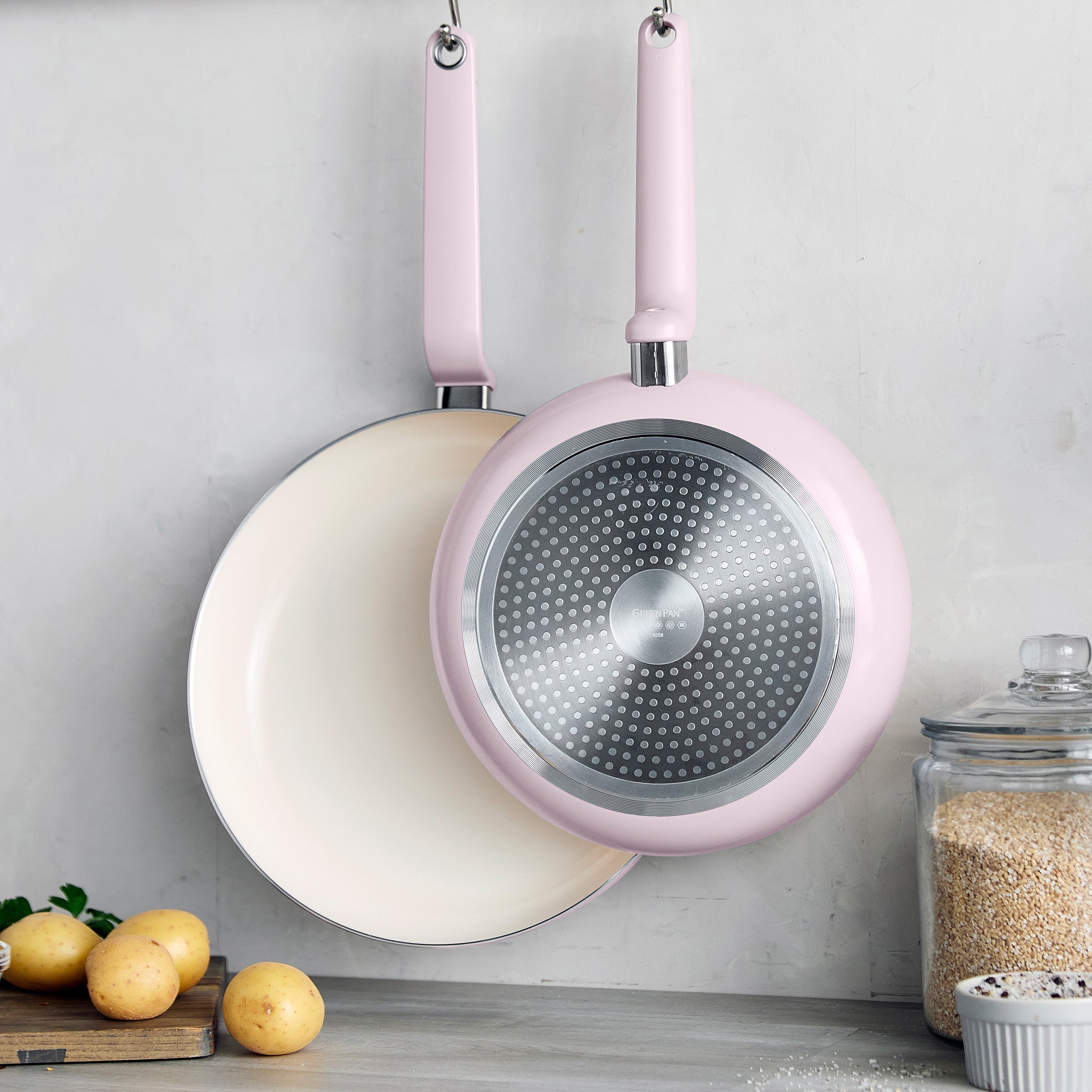 Set of two pink ceramic nonstick frypans hanging above a kitchen counter with potatoes, rice, and a cutting board.