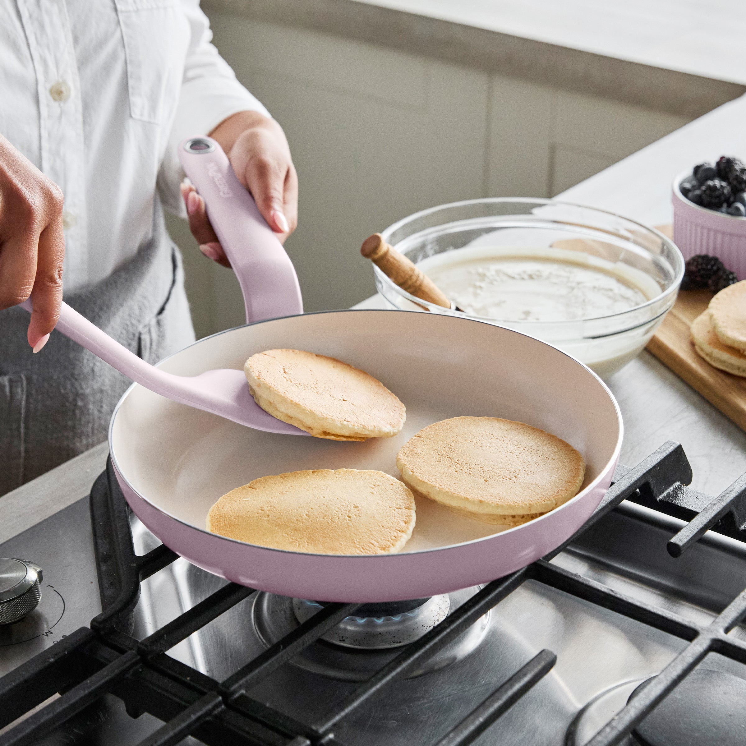 Pancake being flipped with a lavender spatula in a pink ceramic nonstick frypan, batter and berries beside the pan.