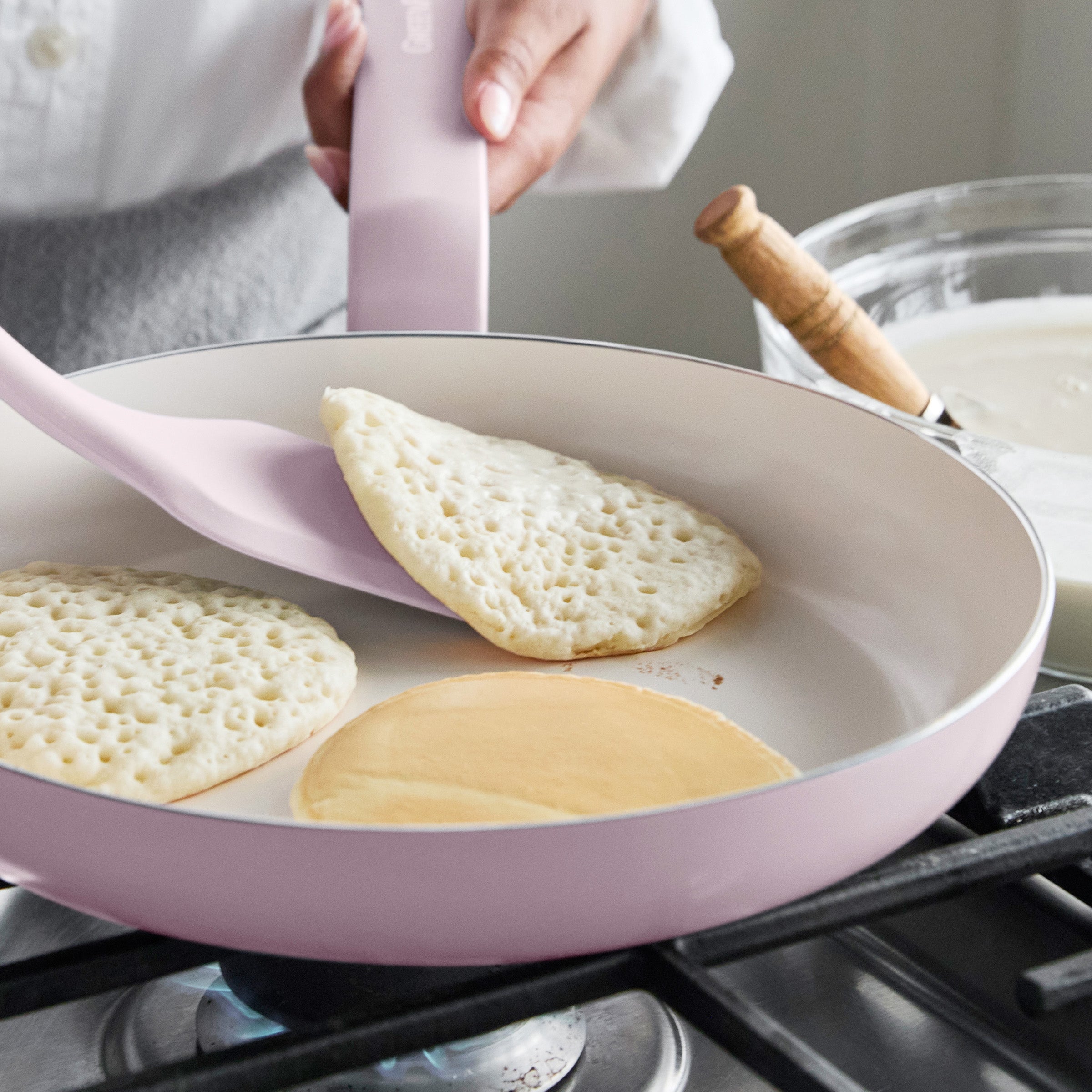 A pancake being flipped in a pink ceramic nonstick frypan, with a batter bowl and whisk on the stove nearby.