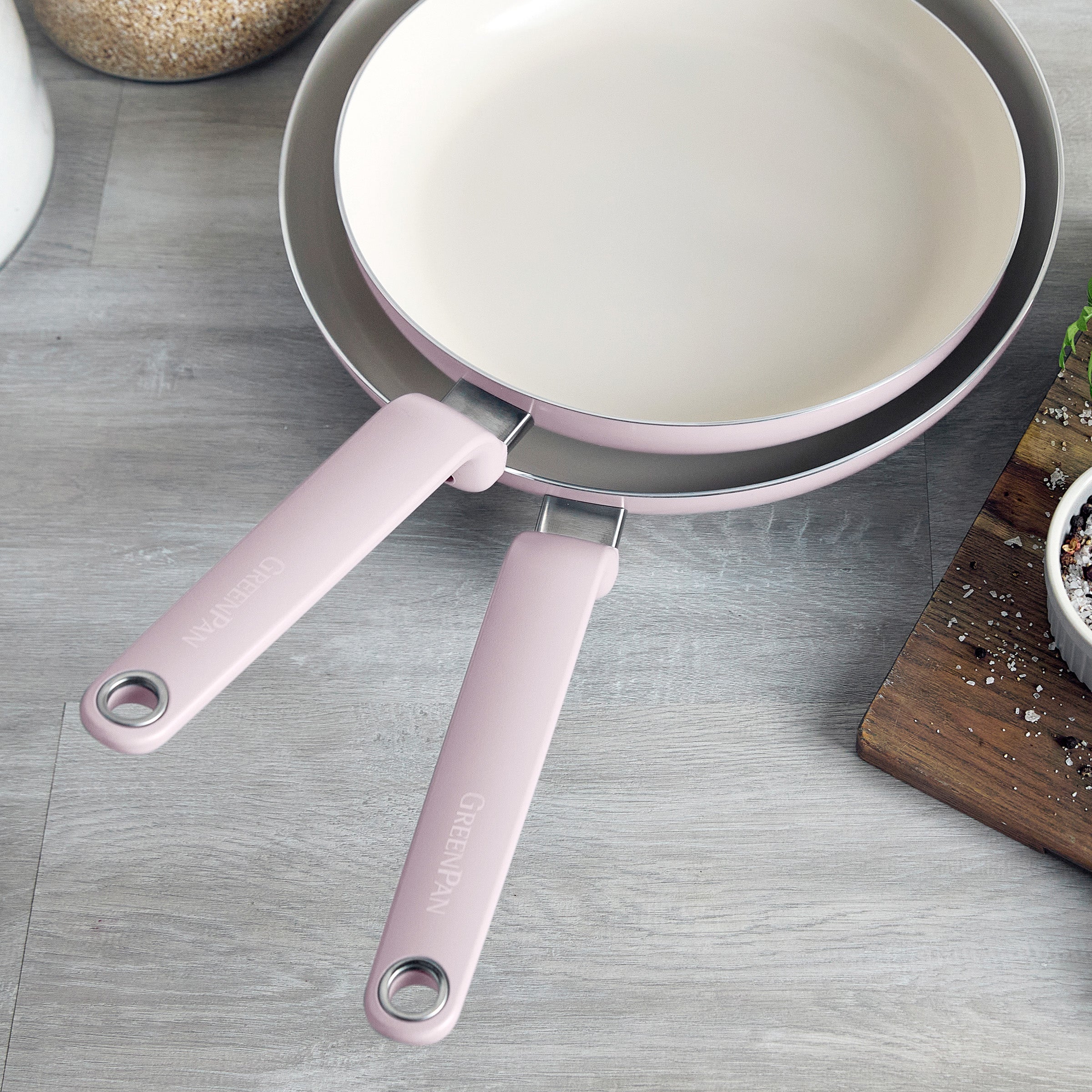 Two pink ceramic nonstick frypans on a grey countertop beside a cutting board with fresh herbs.