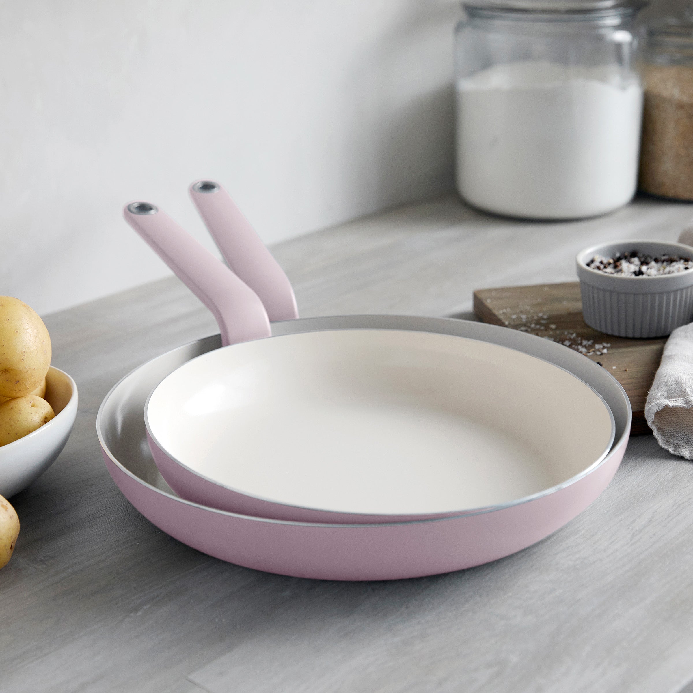 Set of two pink ceramic nonstick frypans, 10 and 12, nested on a kitchen counter with jars, pepper, potatoes, and a cutting board.