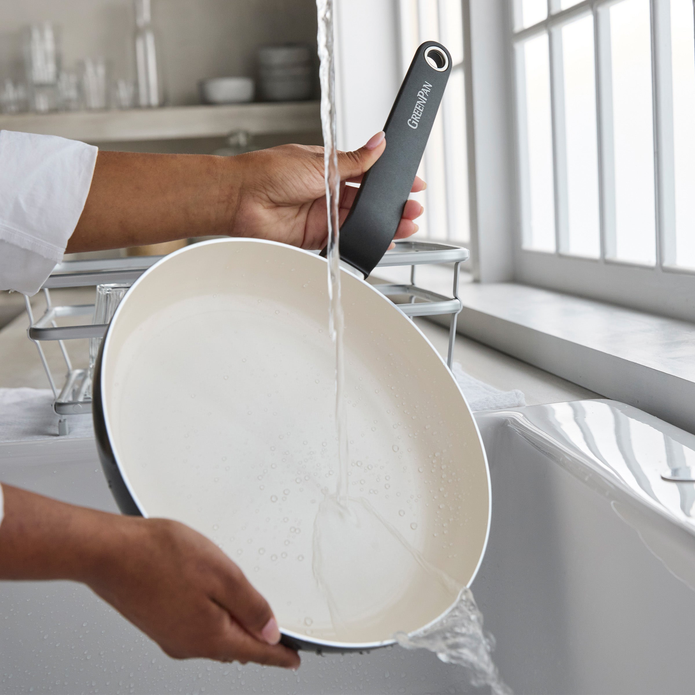 A hand rinses a black ceramic nonstick pan from a 10-piece cookware set under running water in a kitchen sink.