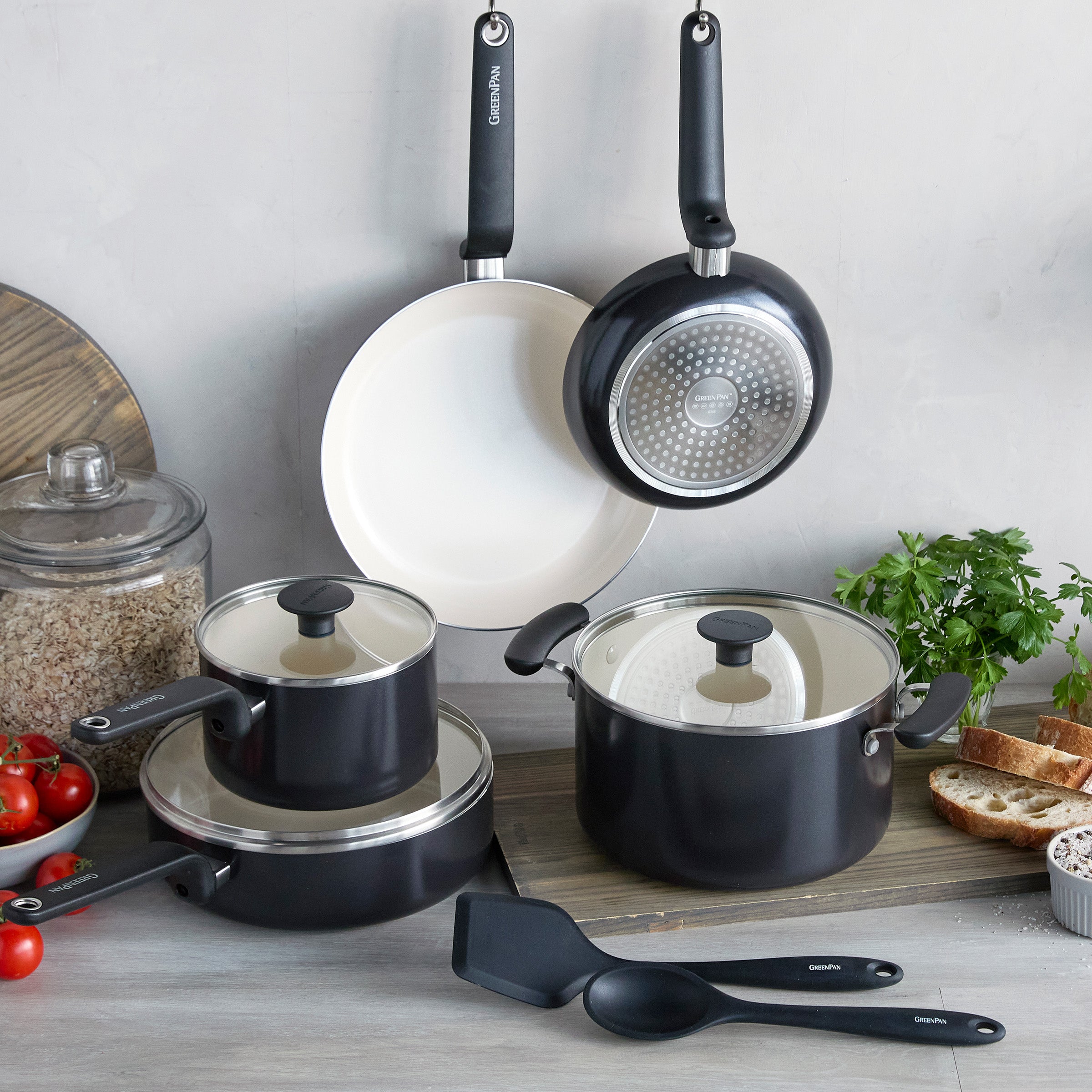 Black 10-piece ceramic nonstick cookware set on a kitchen counter with tomatoes, herbs, bread, and oats nearby.