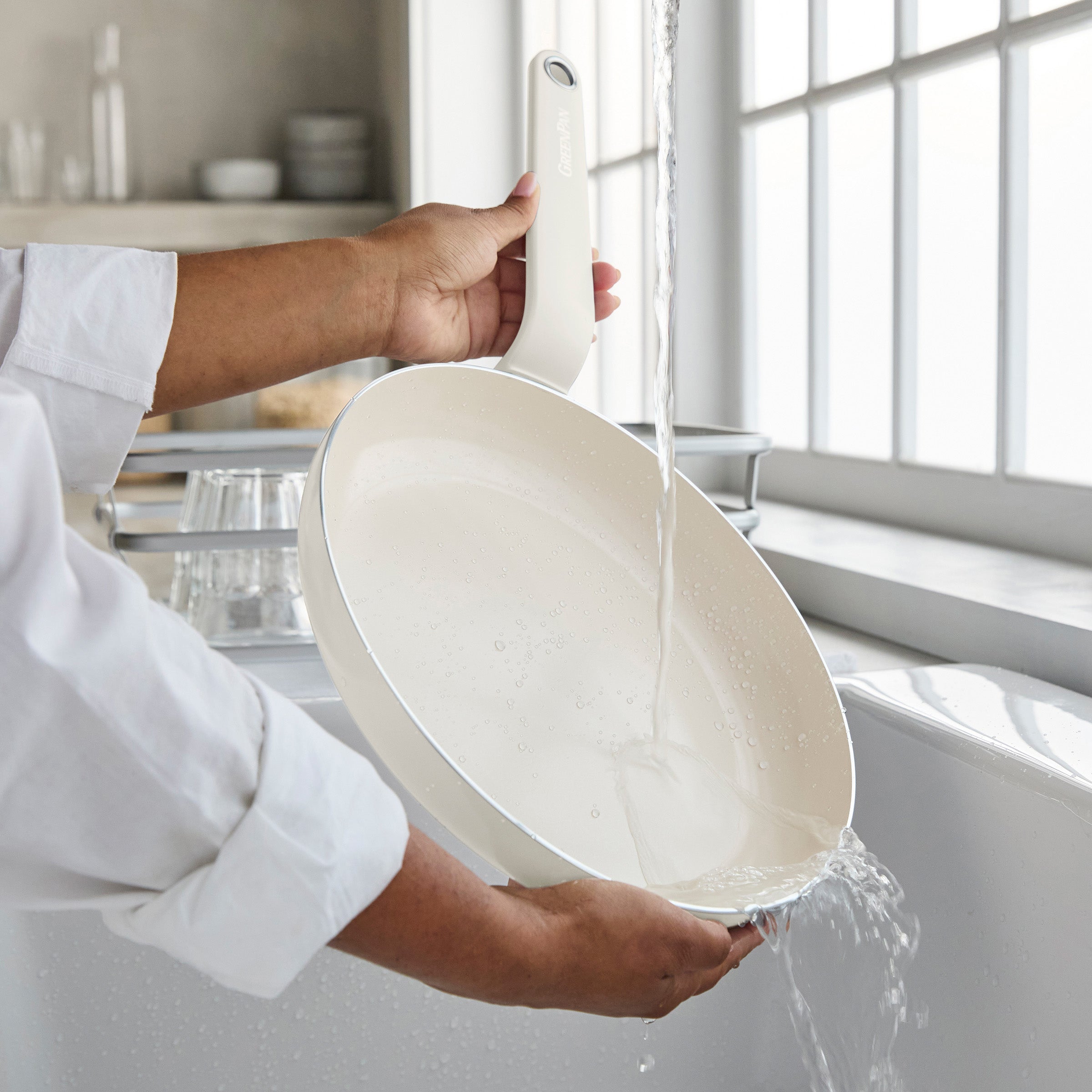 Rinsing a cream ceramic nonstick pan from a 10-piece set under running water in a bright kitchen by a window.