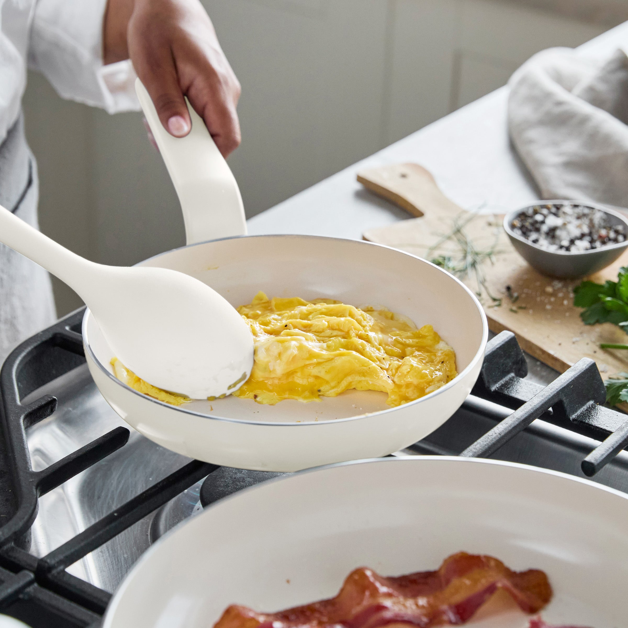 Scrambled eggs cooked with a white spatula in a cream ceramic nonstick pan; bacon pieces are cooking nearby.