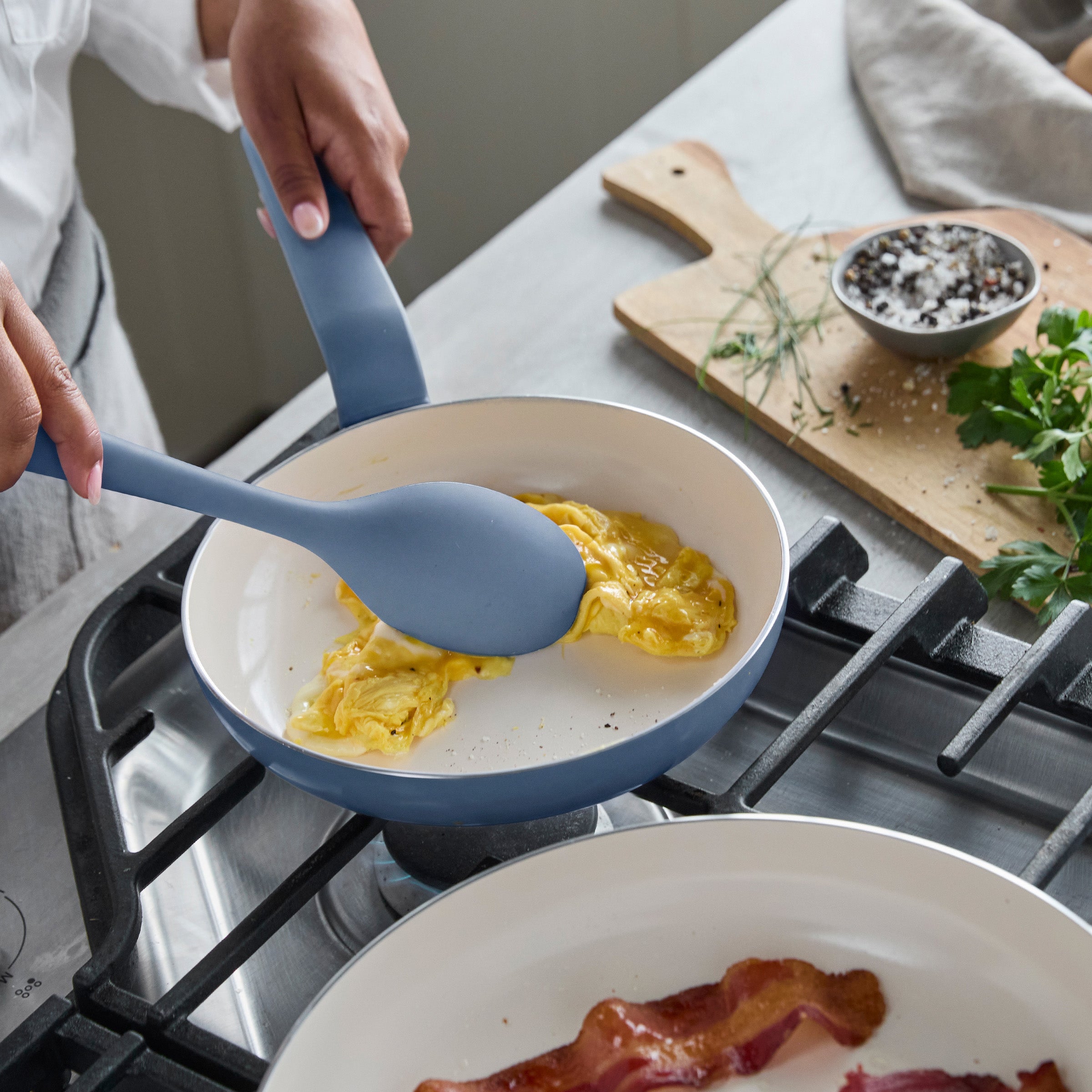 Scrambled eggs cooking in a blue ceramic nonstick pan on the stove, with bacon strips frying in a nearby skillet.