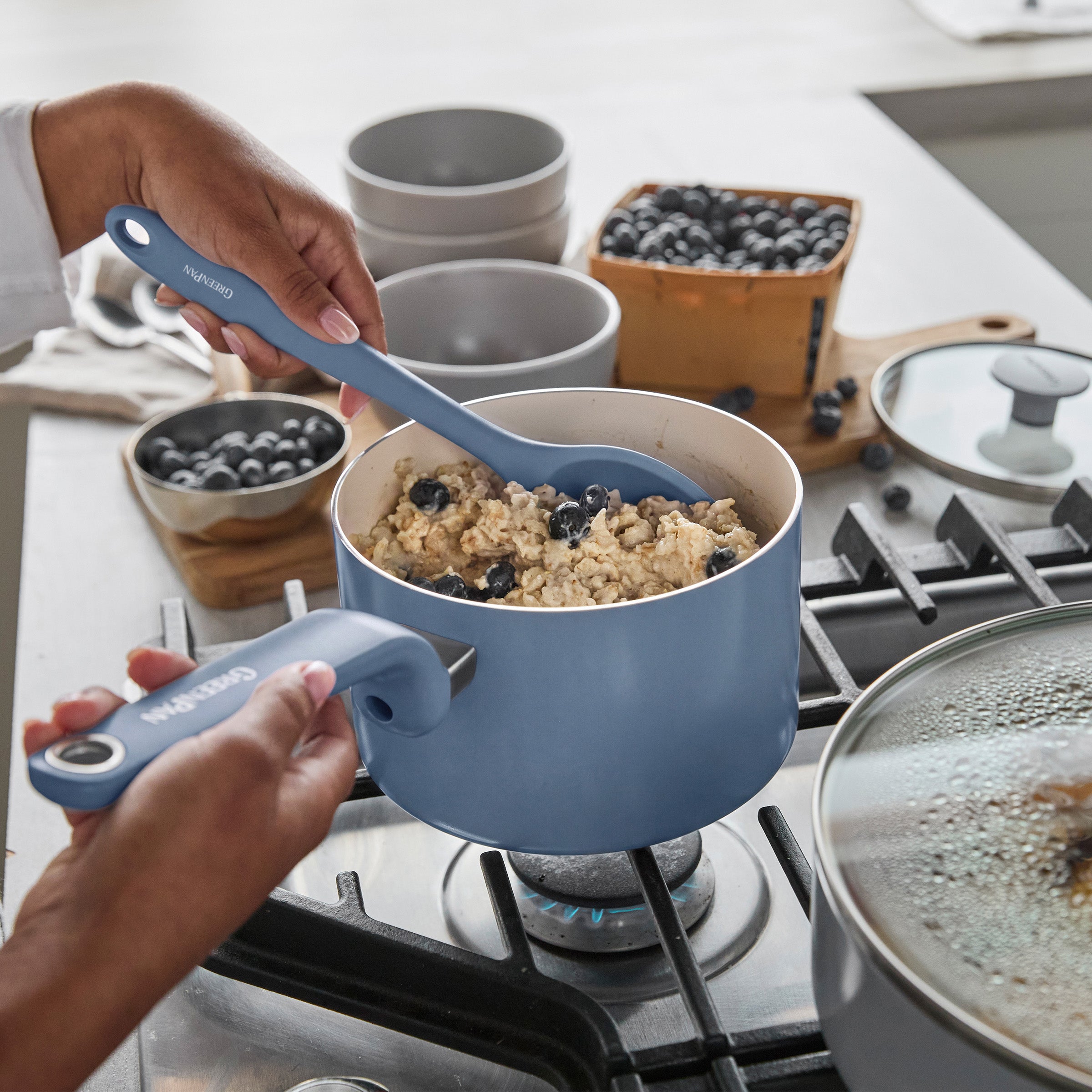Oatmeal with blueberries is stirred in a blue ceramic nonstick pot, with bowls and fresh berries nearby.