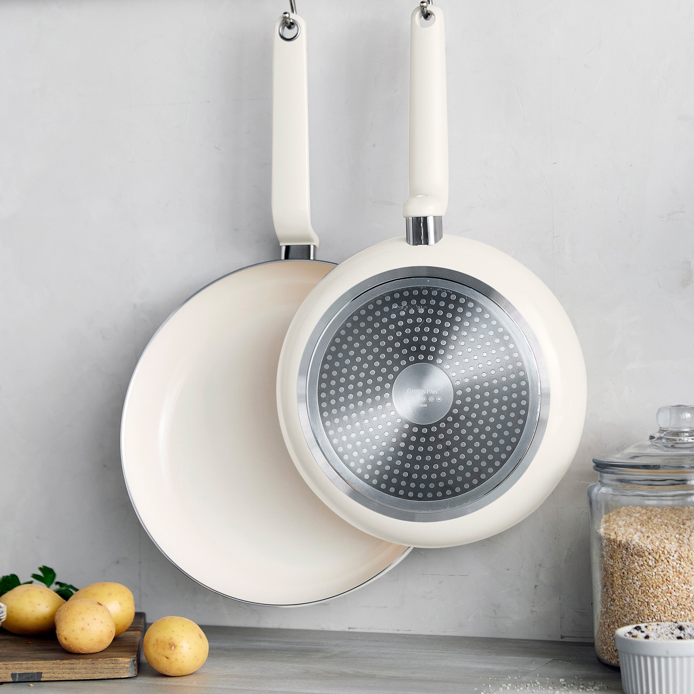 Set of two cream ceramic frypans hanging on a gray wall above potatoes, rice, and a wooden cutting board.