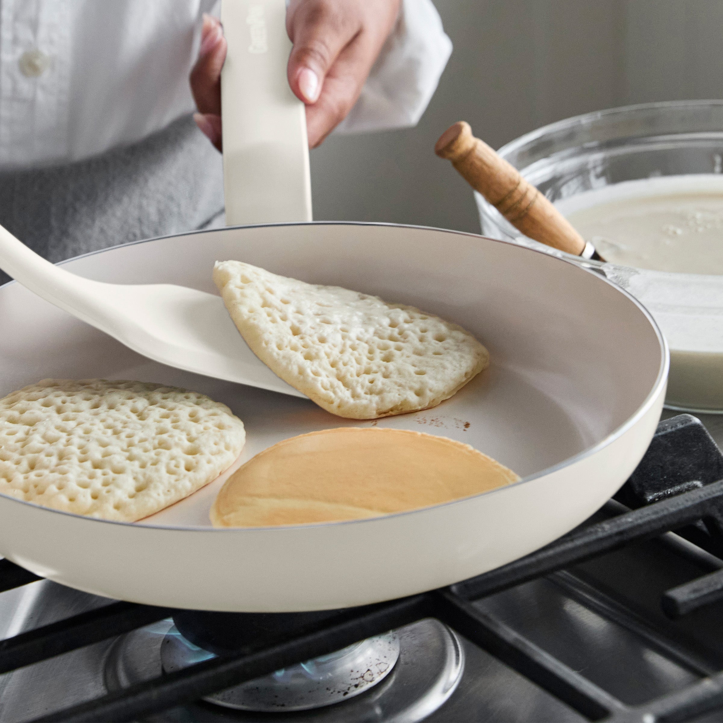 Flipping a pancake with a white spatula in a cream ceramic nonstick frypan, batter bowl on the counter nearby.