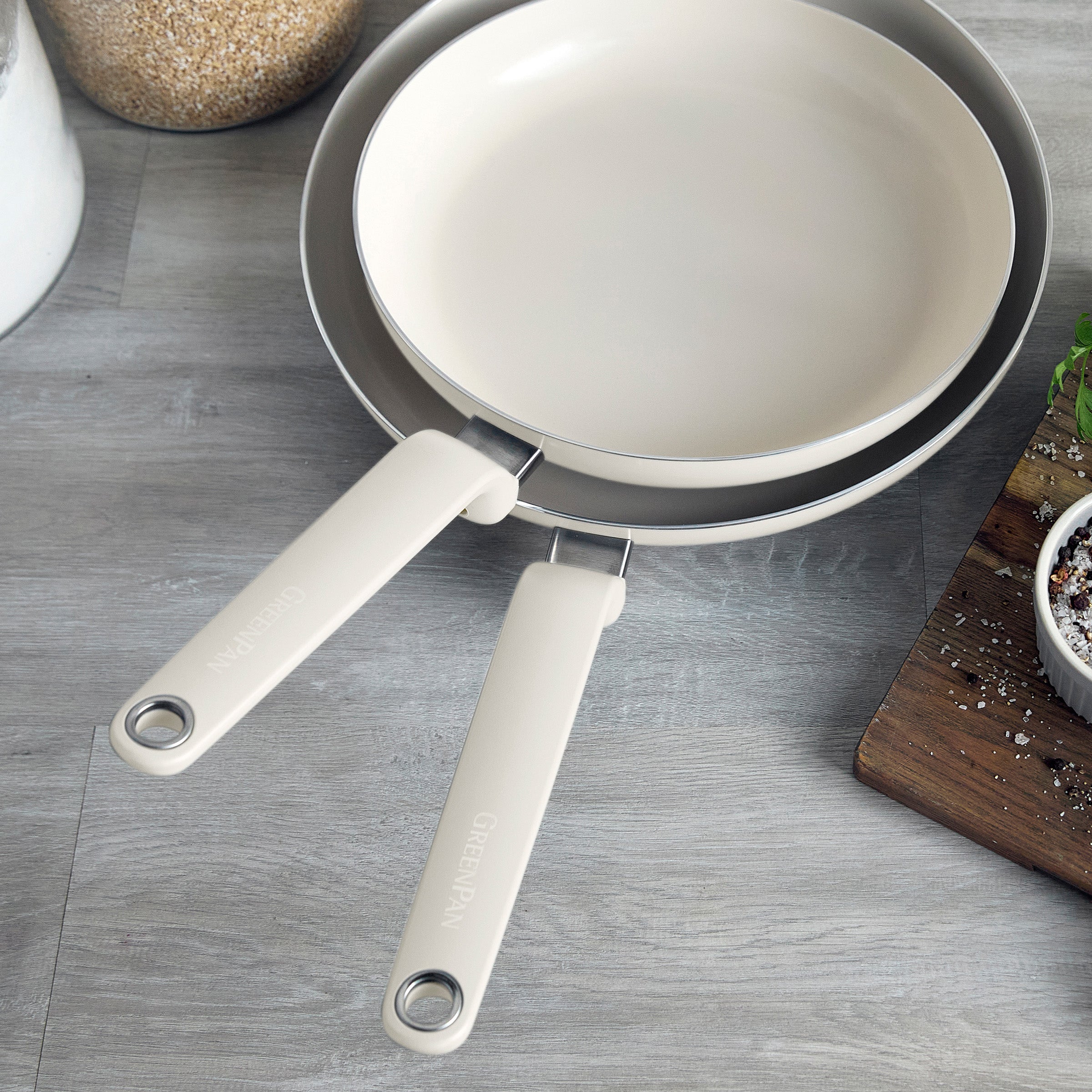 Two cream ceramic nonstick frypans, 10 and 12, on a gray surface beside a cutting board with fresh herbs.
