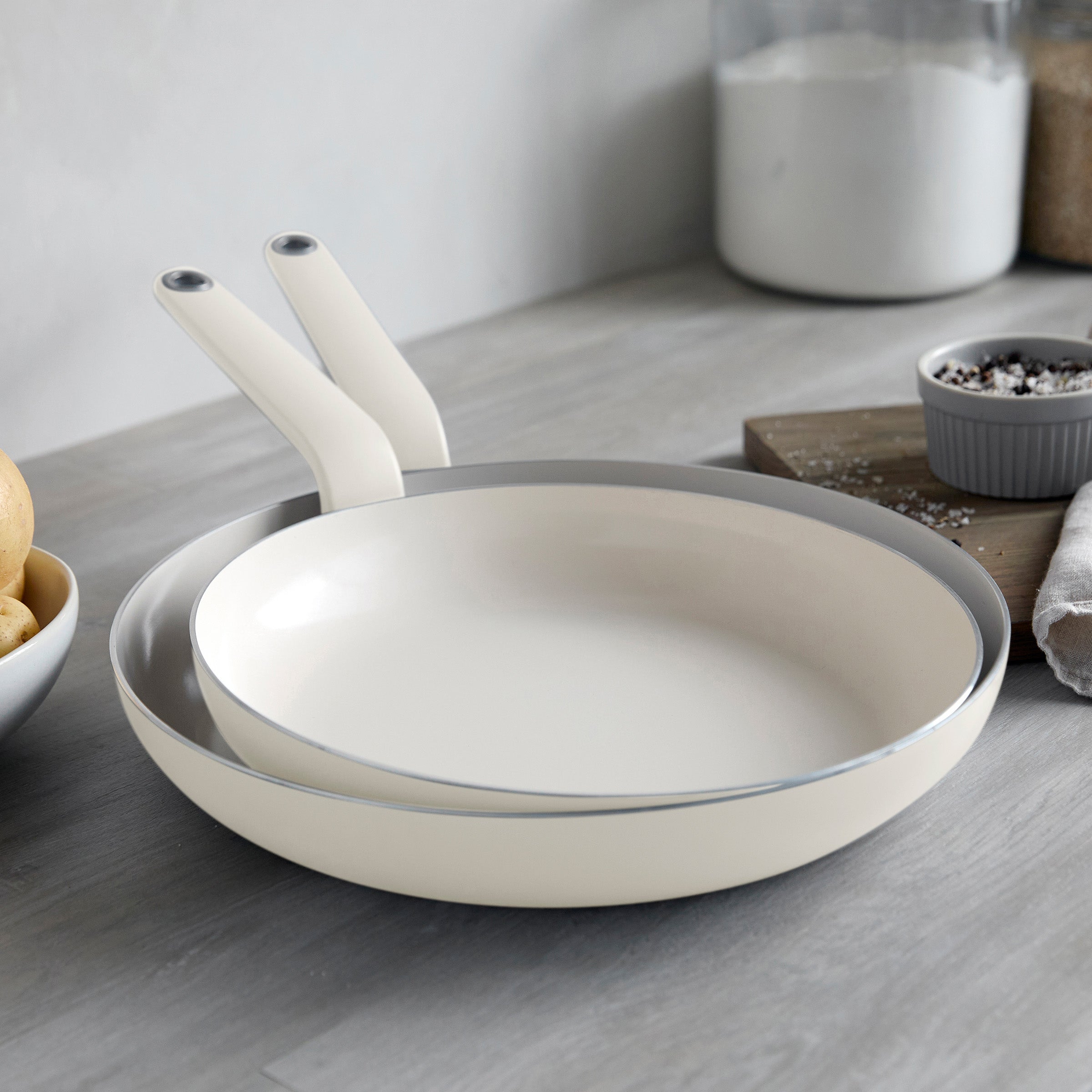 Two cream ceramic nonstick frypans stacked on a kitchen counter with salt, pepper shakers, and a flour jar nearby.