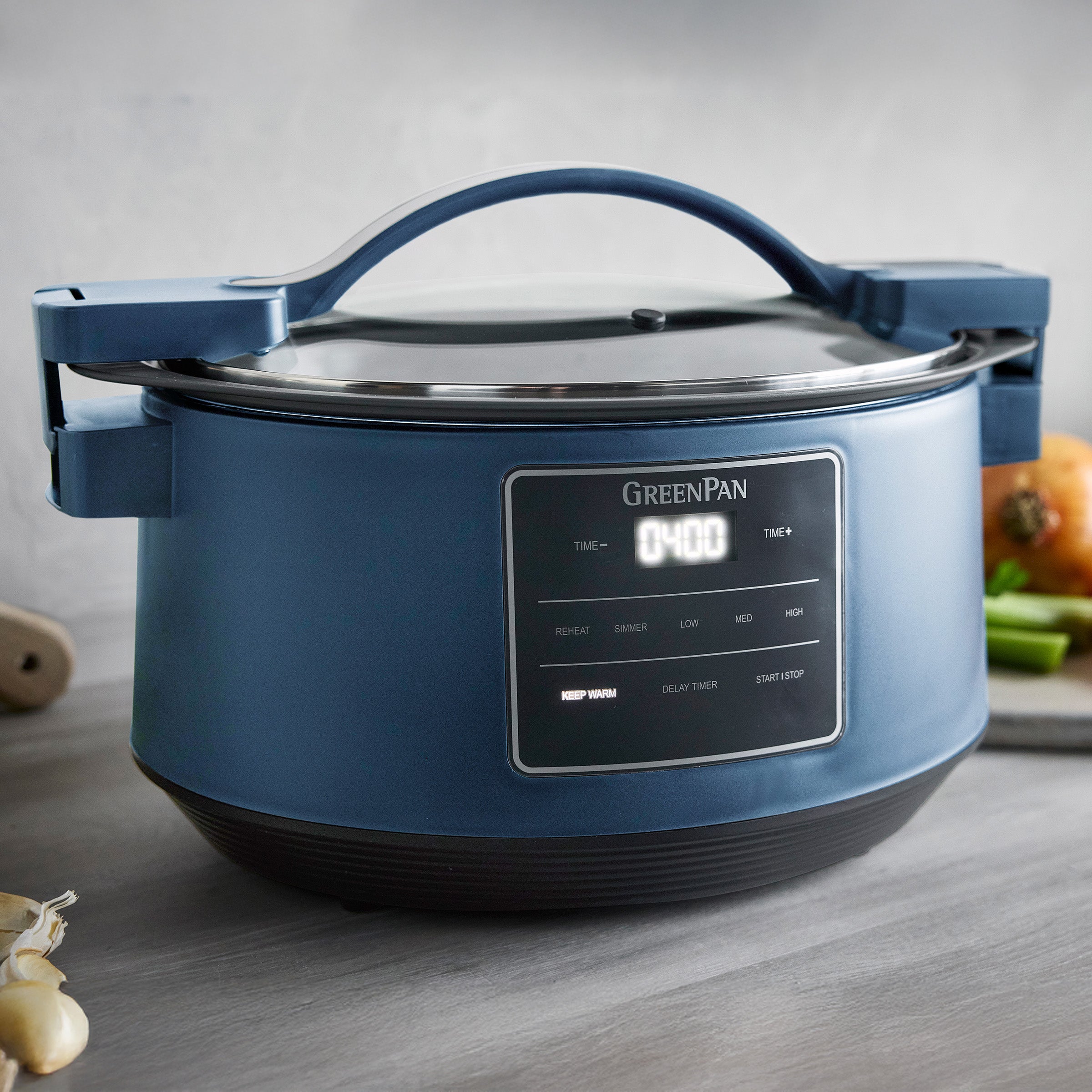 Blue GreenPan slow cooker on a wooden surface with vegetables in the background