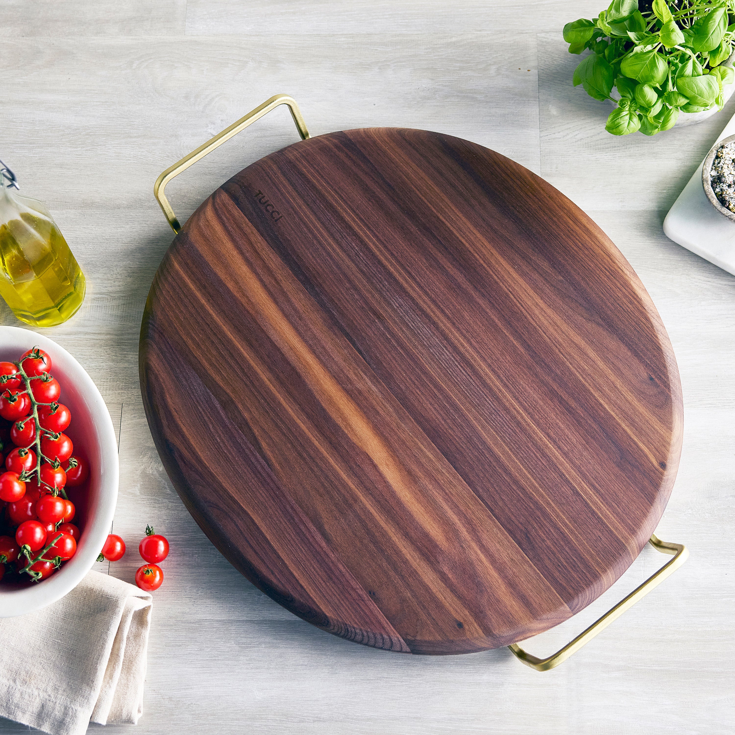 Wooden pizza stone with a handle on a light surface with vegetables and a bottle of oil.