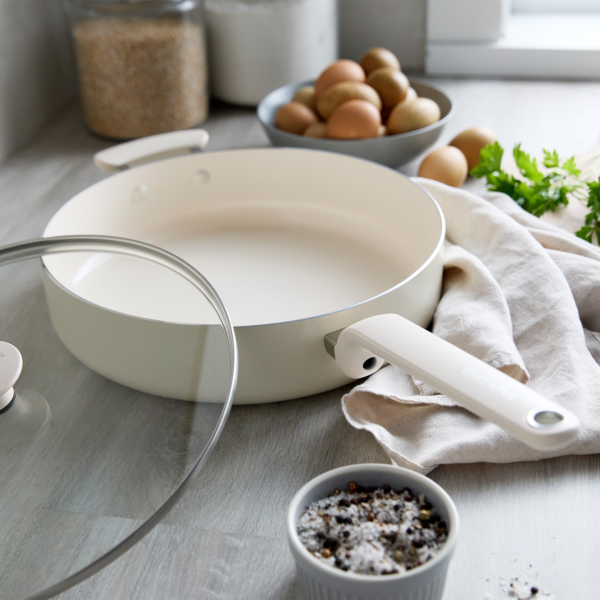 Cream ceramic sauté pan with lid on a kitchen counter, surrounded by eggs, fresh herbs, and black pepper.