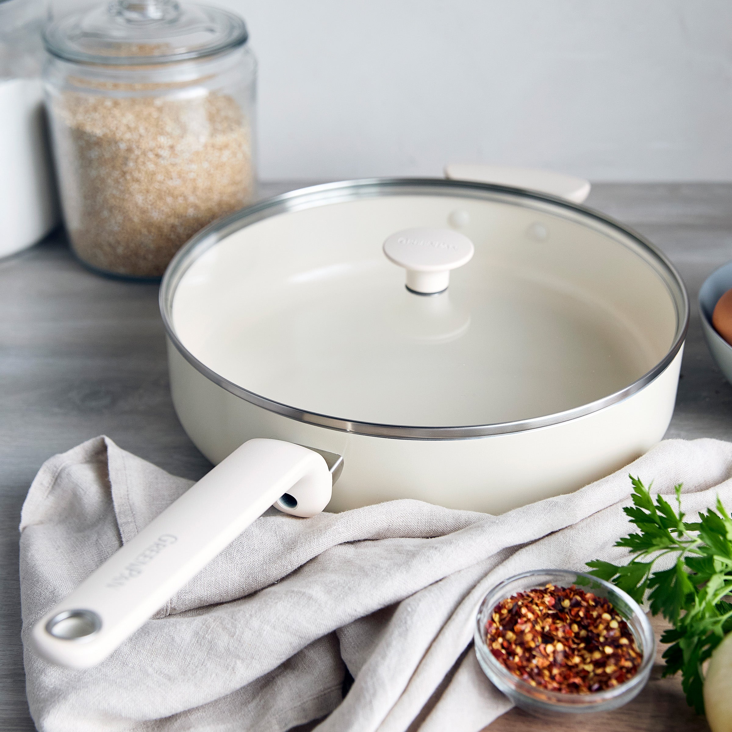 Cream ceramic sauté pan with lid on a cloth, with small bowls of spices and grains around it.