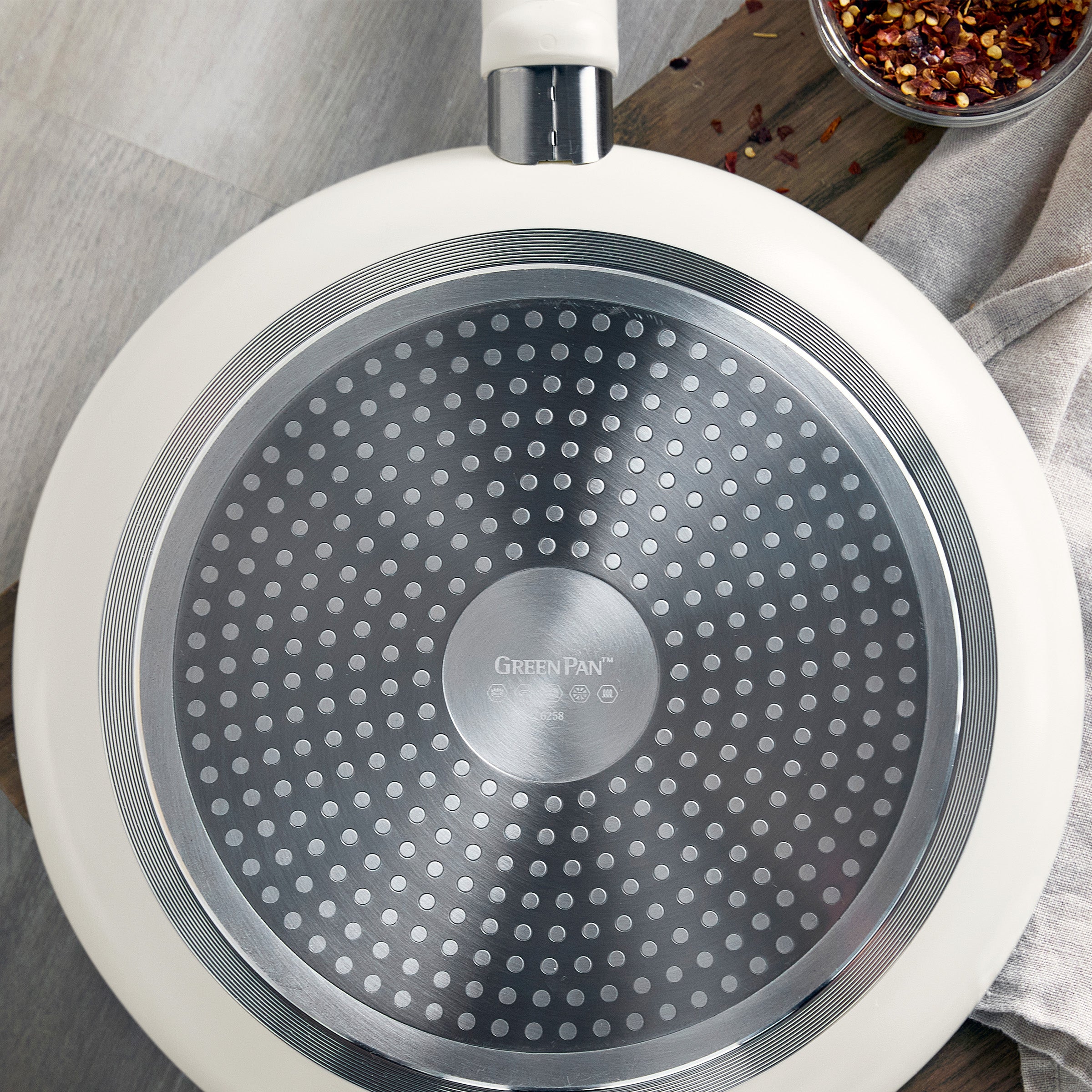 Cream ceramic 5-quart sauté pan with a silver textured base on wood, chili flakes and a towel nearby.