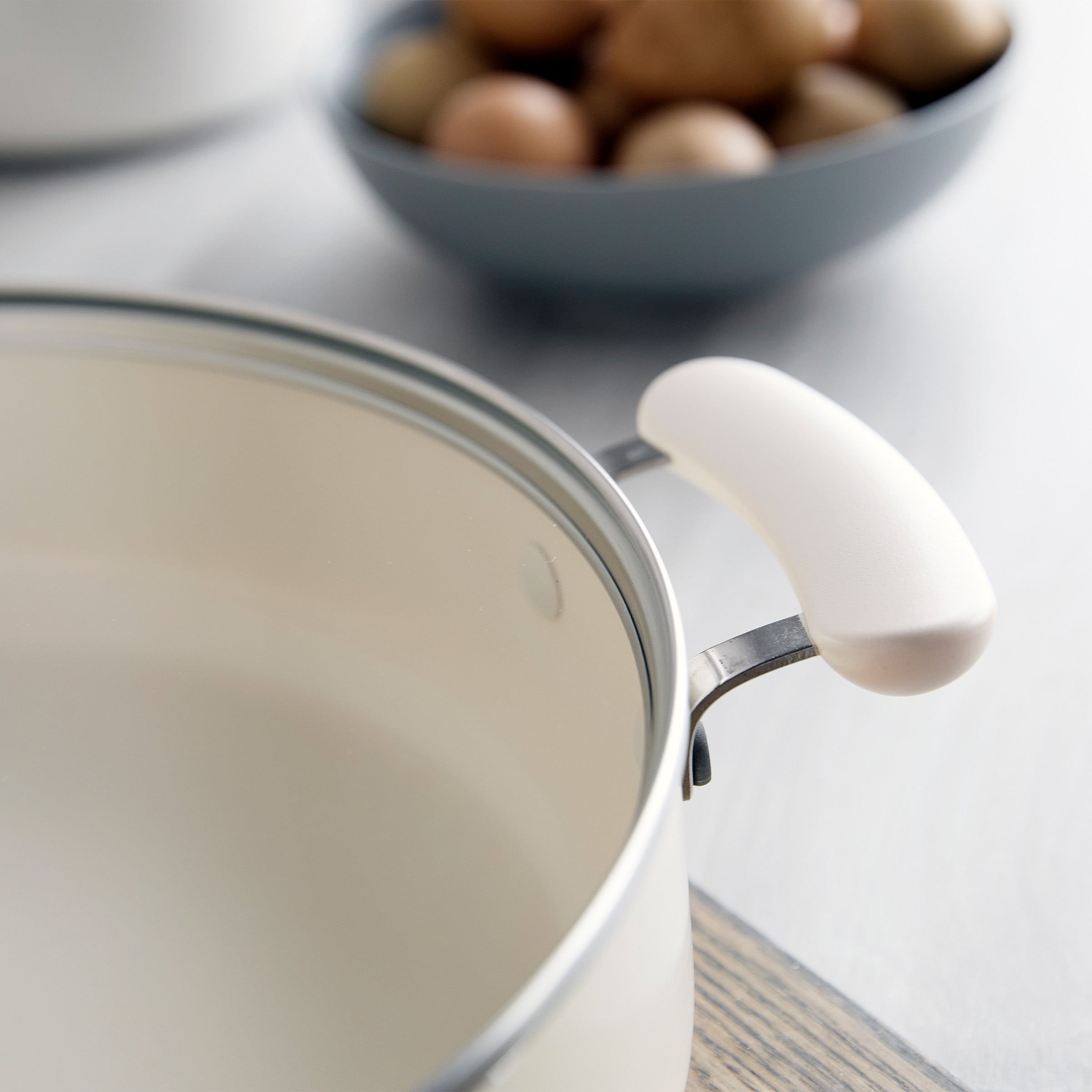 Cream ceramic sauté pan with lid and handle, Rio Advanced model; blurred potatoes are visible in the background.