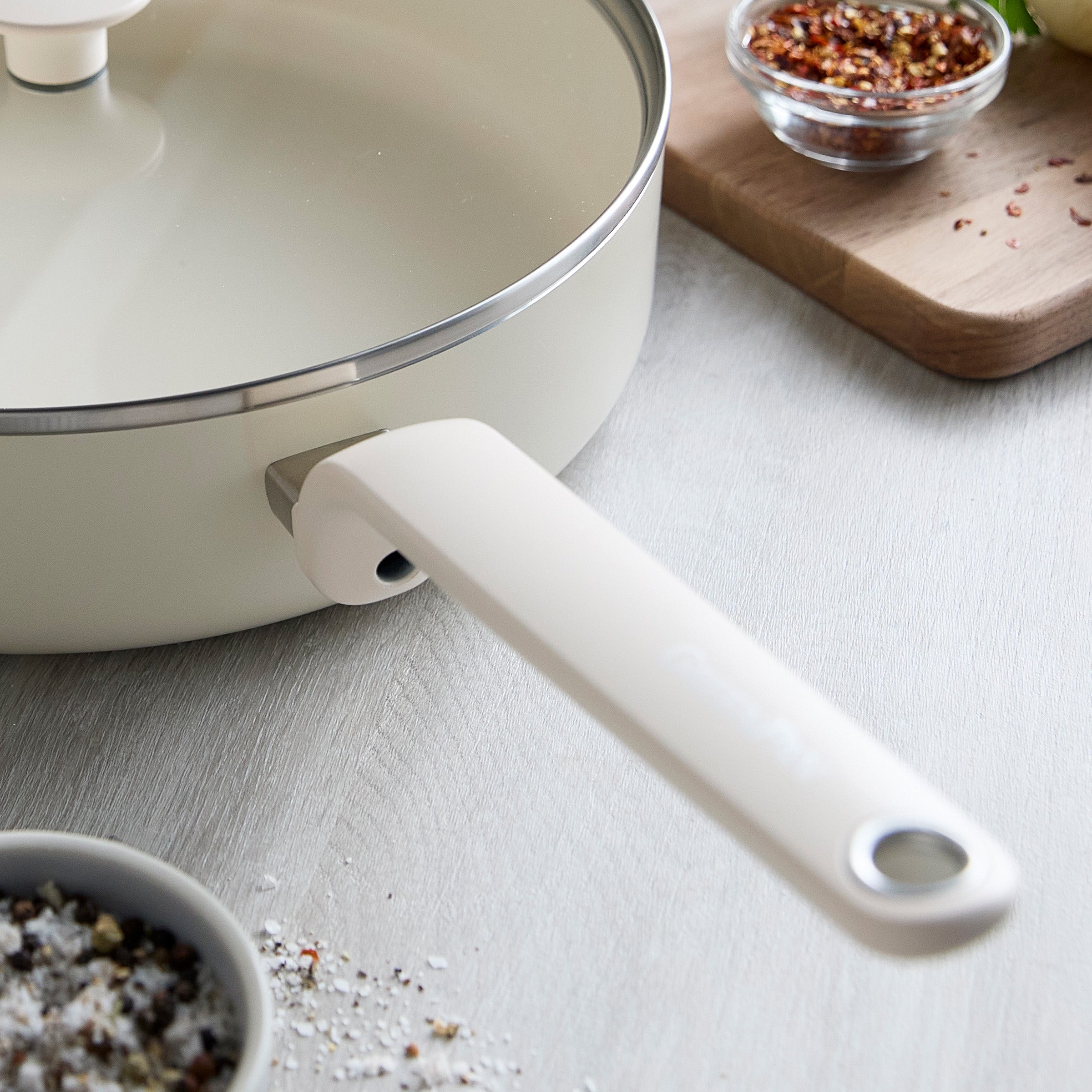 Cream ceramic nonstick sauté pan with lid on counter, next to a spice bowl and cutting board with sliced onions.