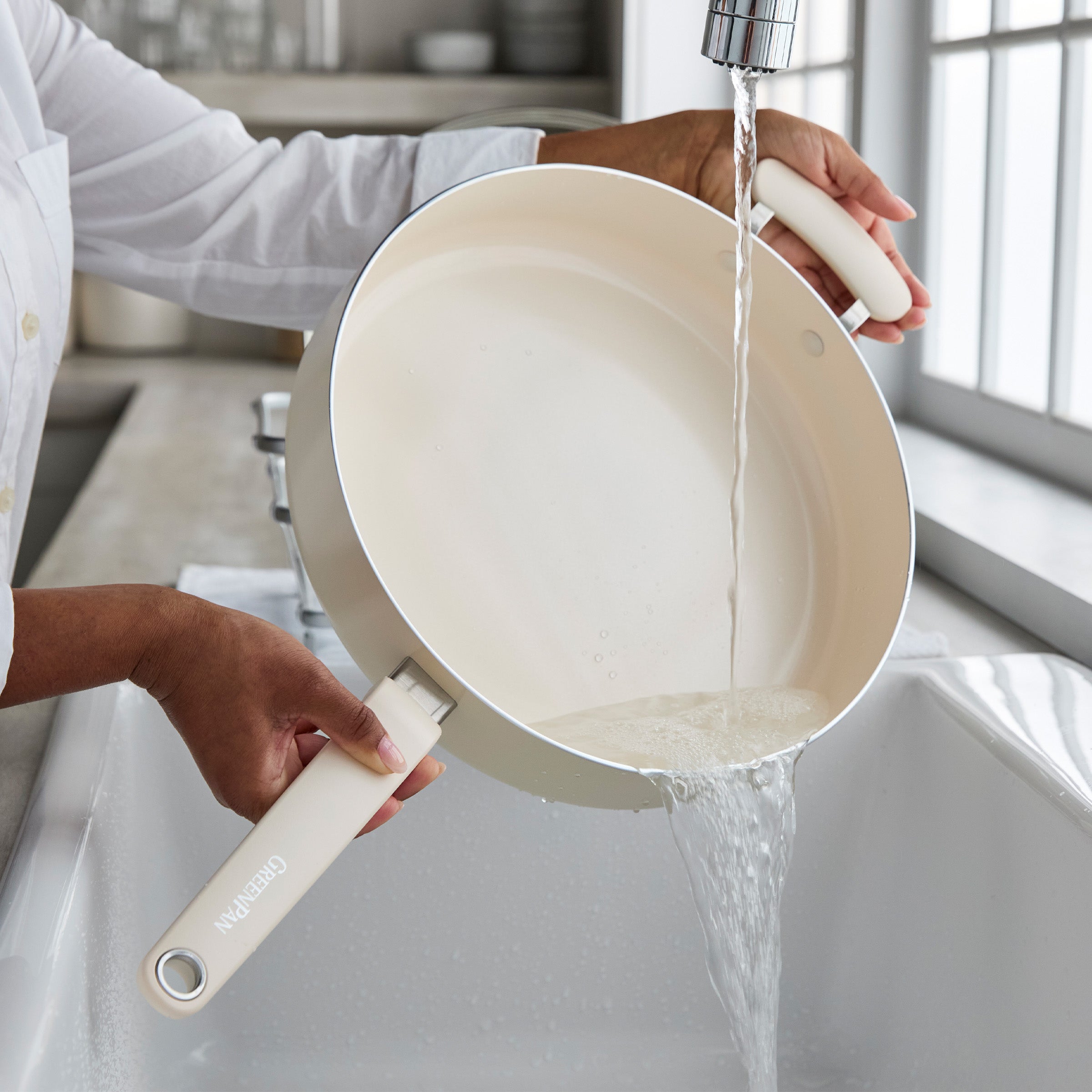 Cream ceramic nonstick sauté pan with lid being rinsed under water in a bright kitchen sink.