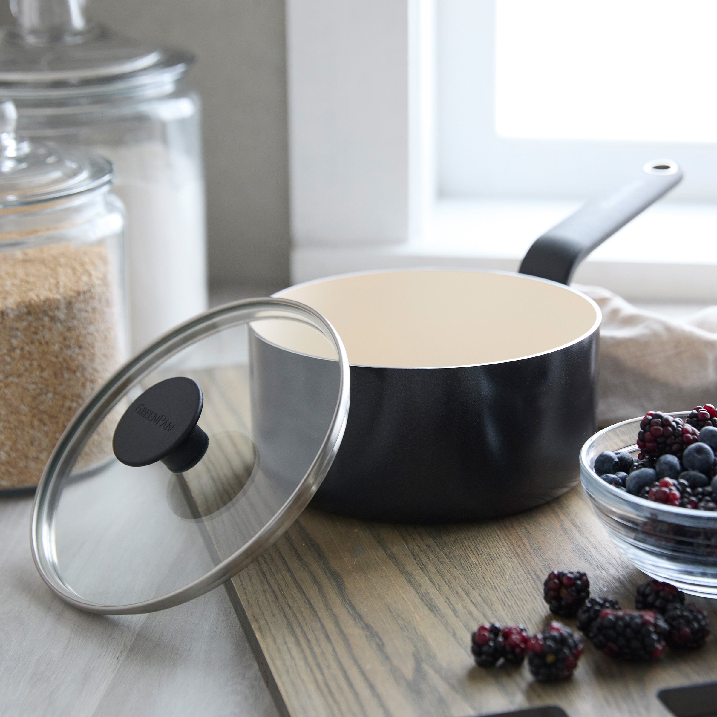 Black ceramic 2-quart saucepan with lid, open on a wooden surface beside sugar jars and a bowl of berries.
