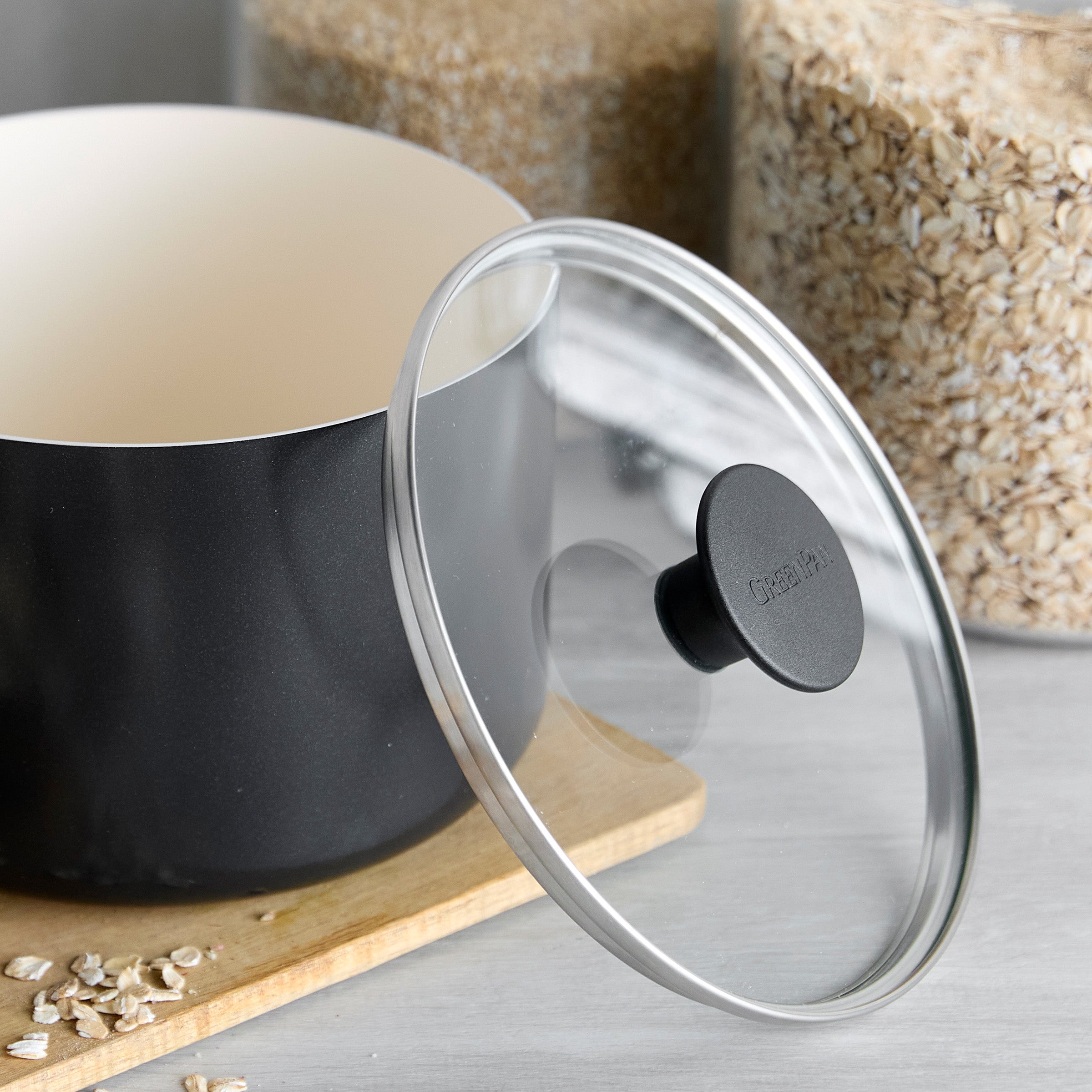 Black ceramic 2-quart saucepan with lid on a wooden board, surrounded by jars of grains and scattered oats.