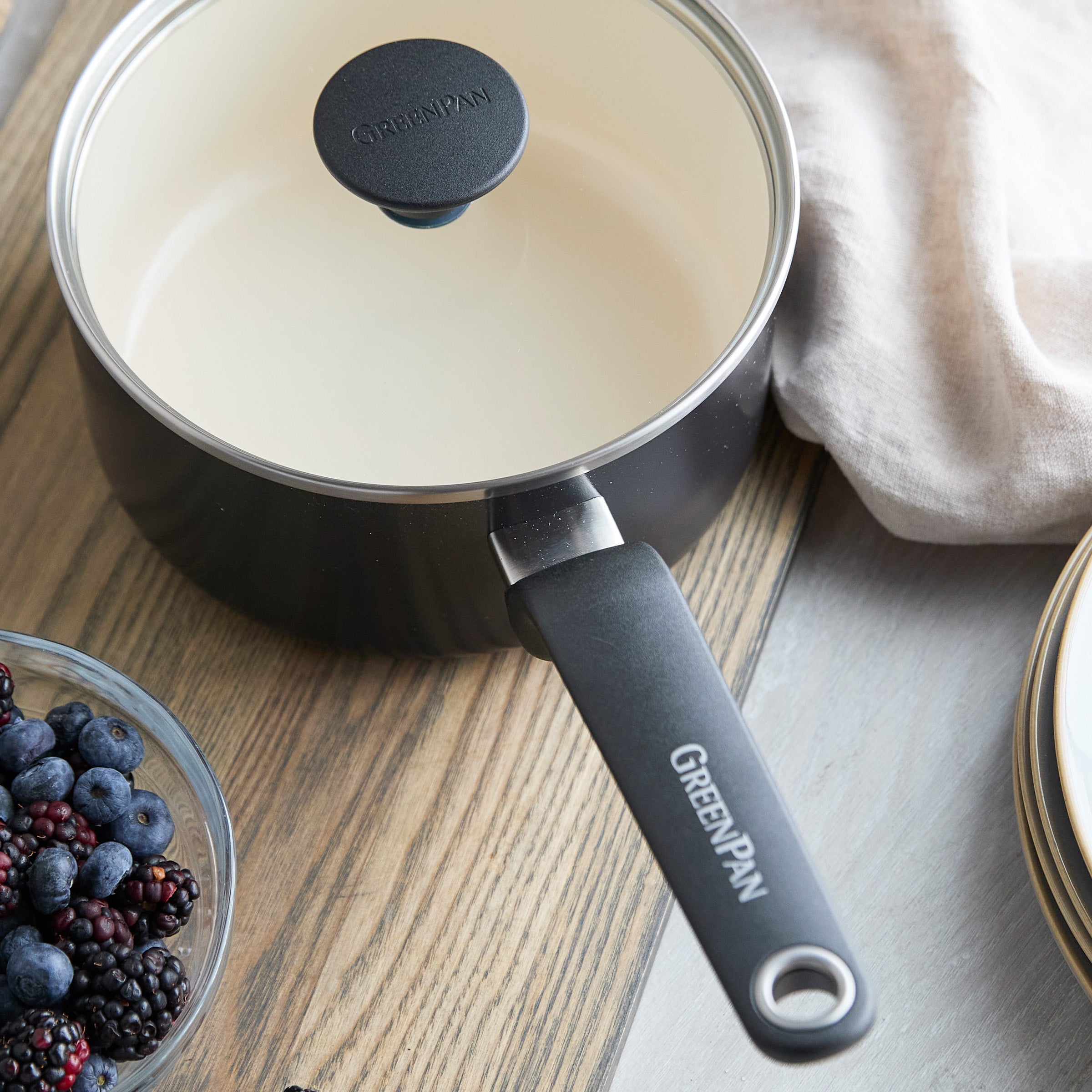 Black ceramic 2-quart saucepan with lid on a wooden table, next to plates, beige cloth, and berries.