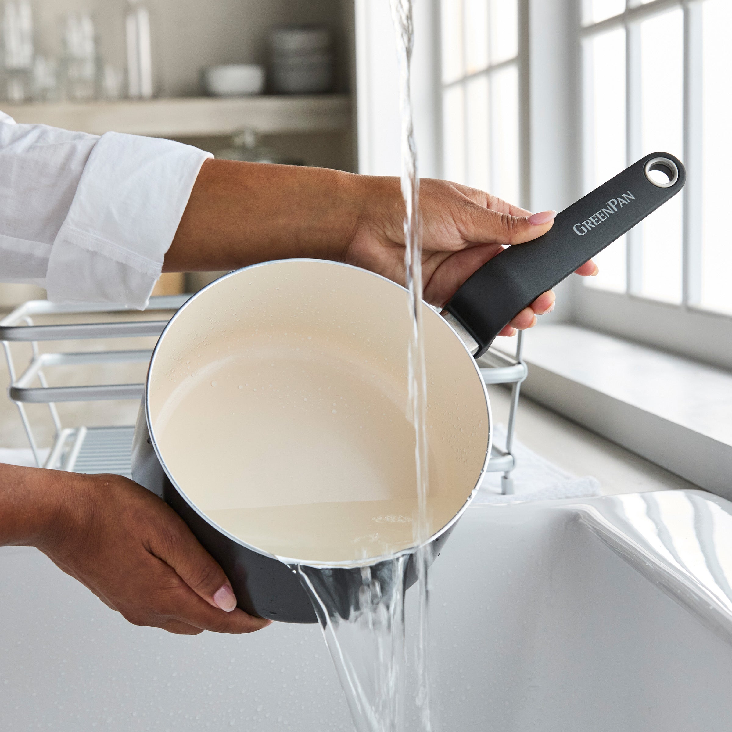 A black ceramic 2-quart saucepan with lid being rinsed under running water at a kitchen sink near a window.