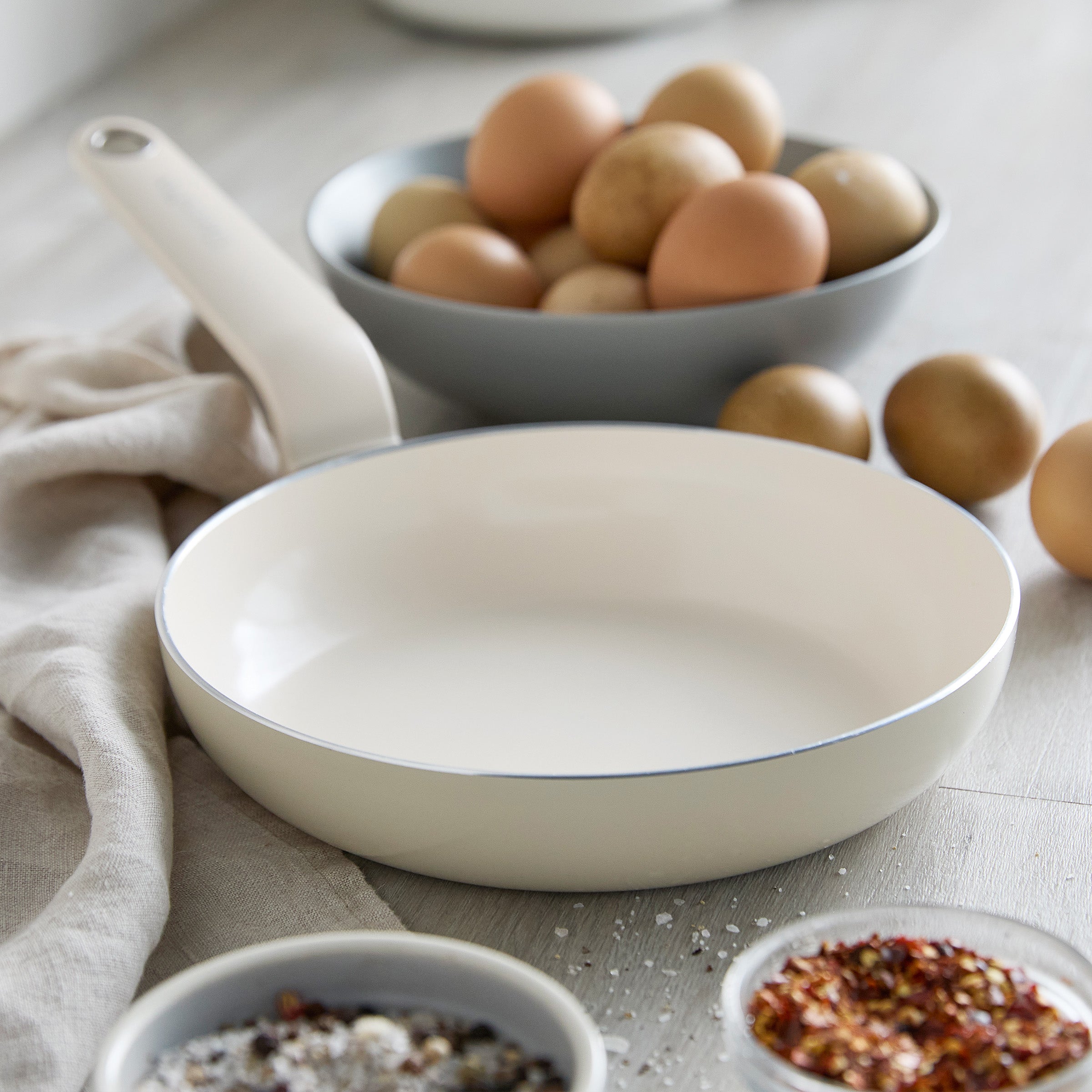 Cream ceramic nonstick 8-inch frypan on a tabletop near bowls of eggs, spices, and a beige cloth.