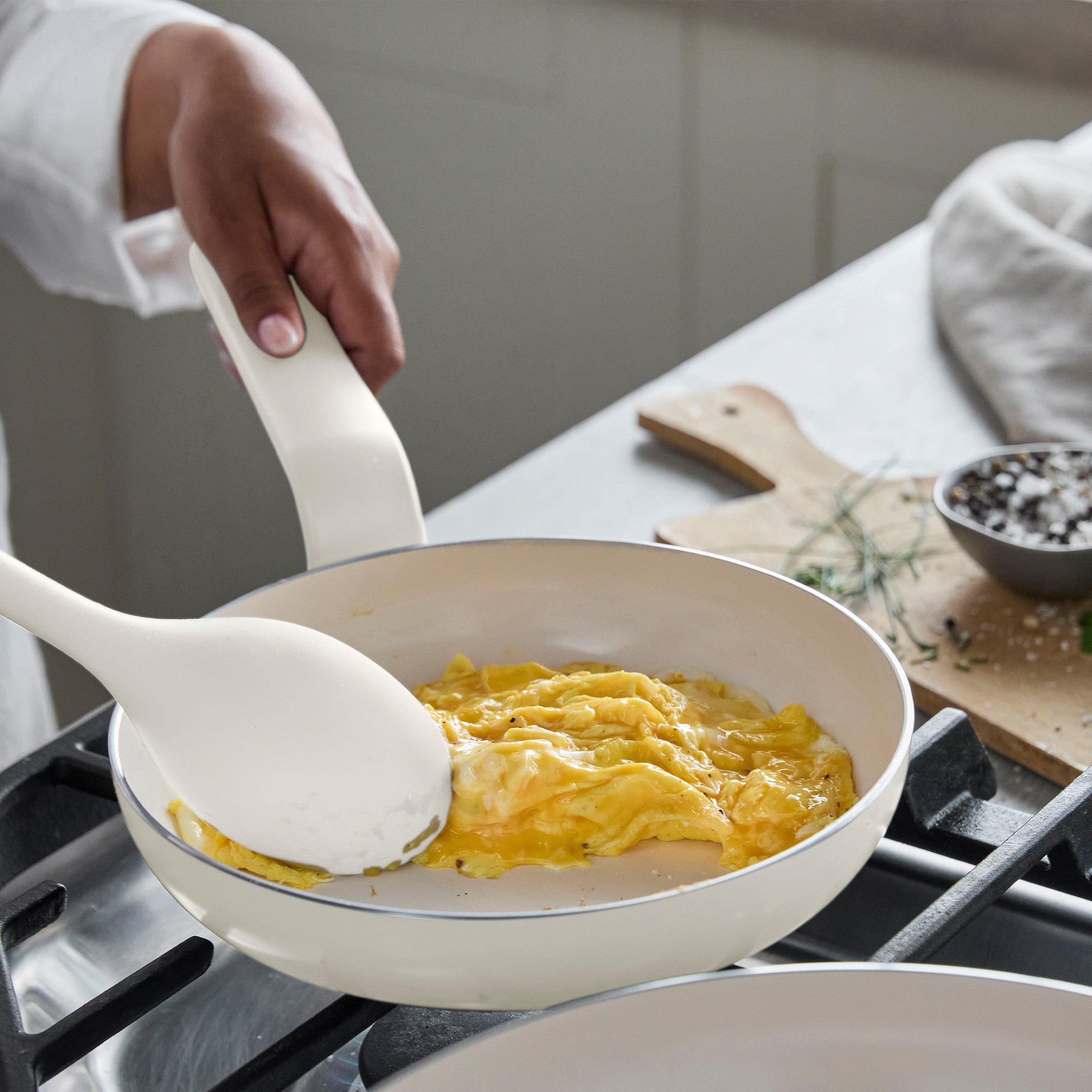 Scrambled eggs being stirred in a cream-colored ceramic nonstick 8-inch frypan on a stovetop with a spatula.