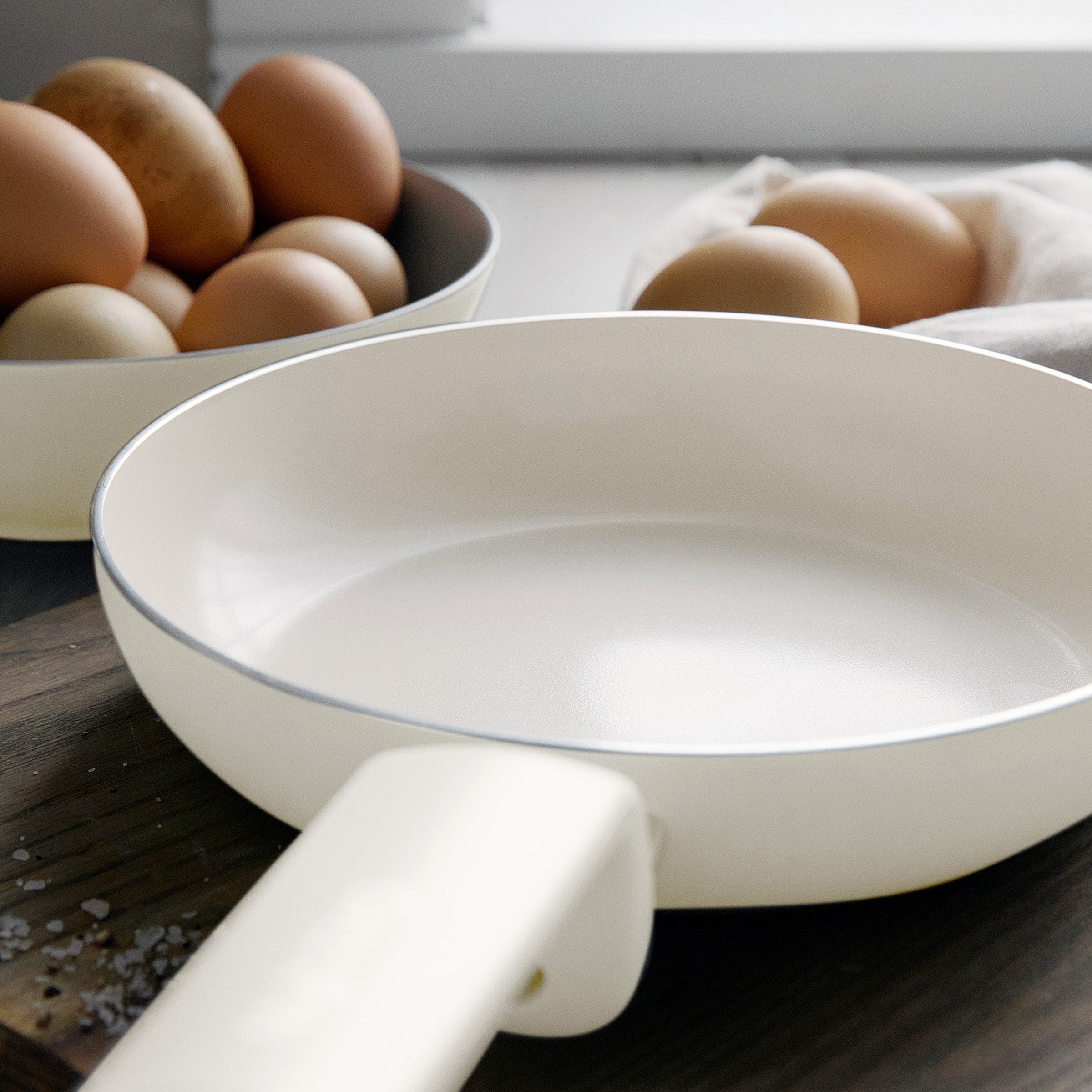 Cream ceramic 8-inch frypan on a wooden surface, with brown eggs in a bowl and some on a cloth in the background.