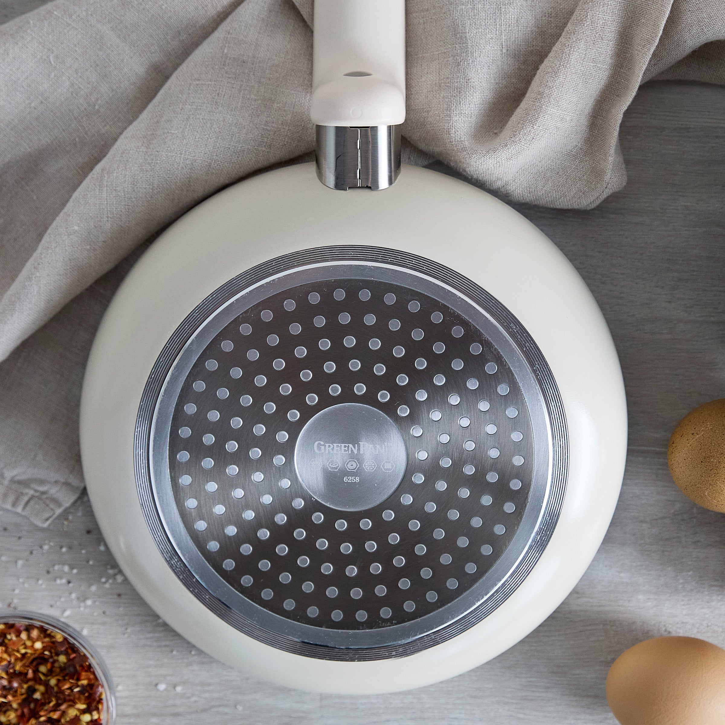 Cream ceramic 8 frypan shown upside down, with a towel, eggs, and spice jars nearby on the surface.