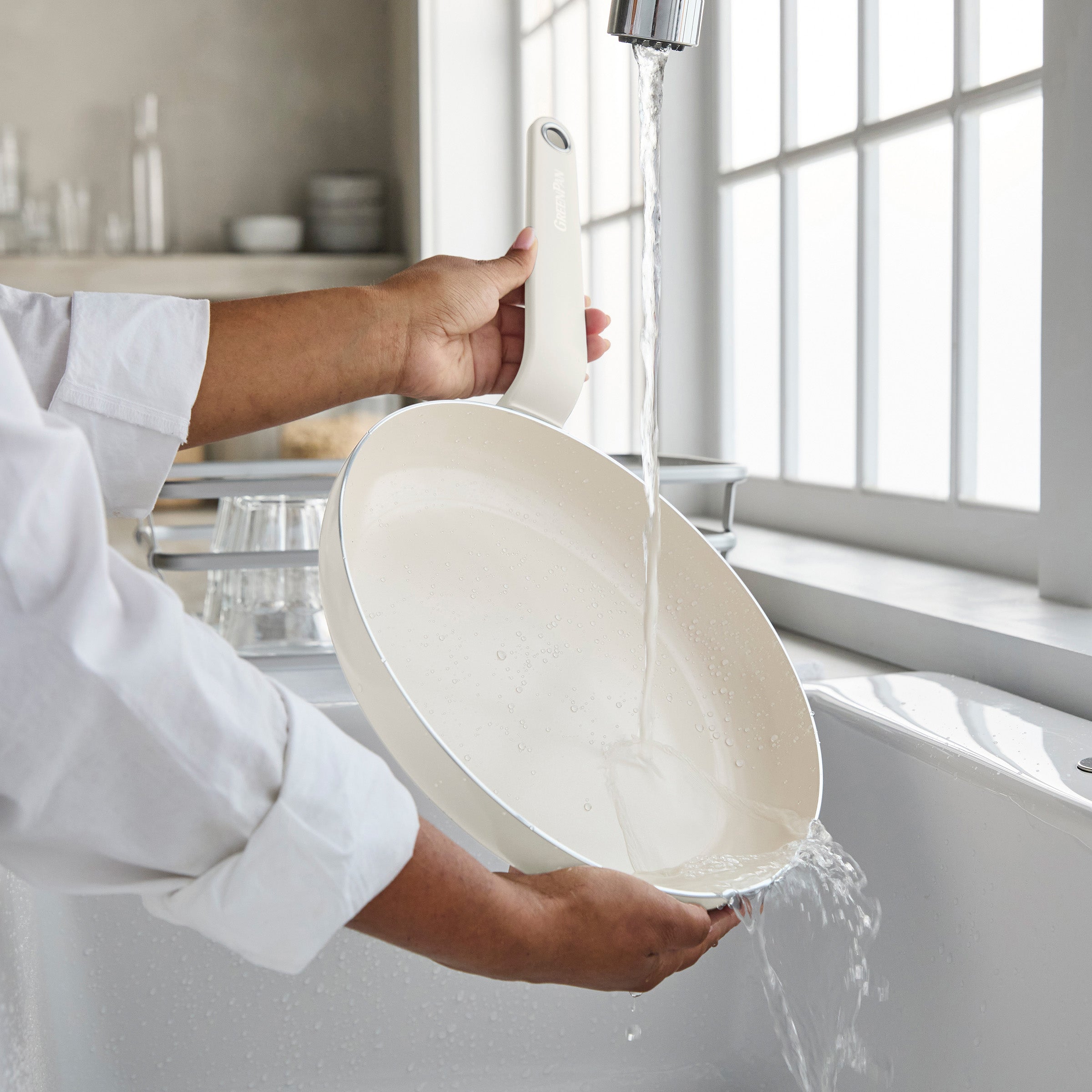 Cream ceramic nonstick 8 frypan being washed under running water in a bright kitchen sink.