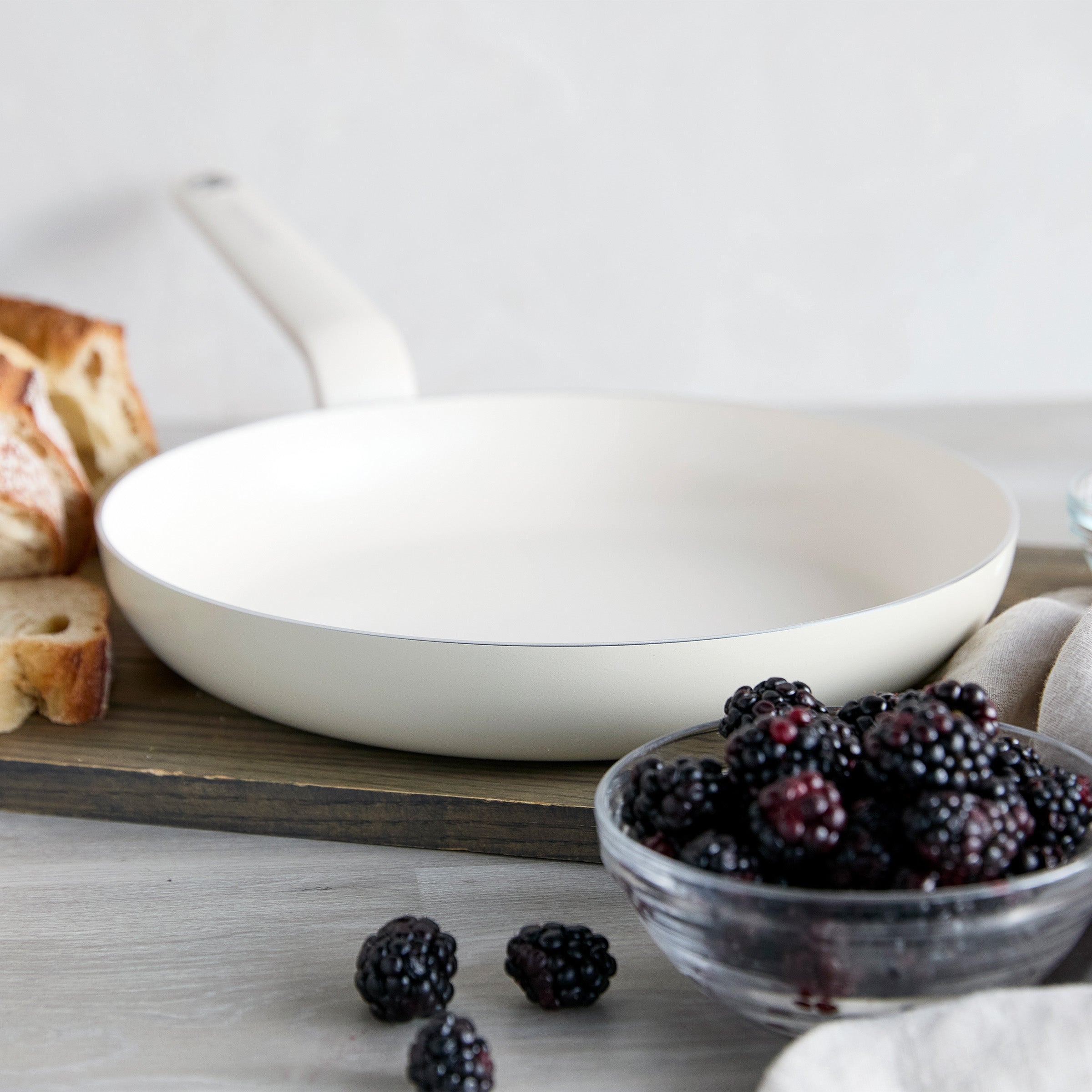 Cream ceramic nonstick 12 frypan on wooden board with sliced bread and a bowl of blackberries nearby.